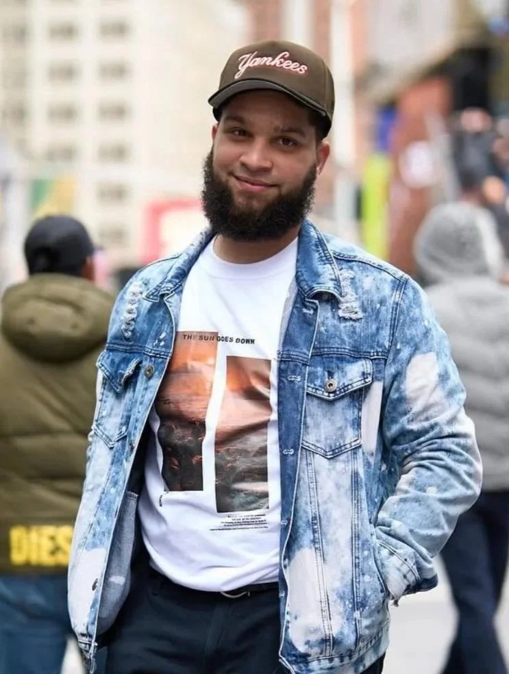 Hombre joven con barba, usando una gorra negra con la palabra 'Yankees', camiseta blanca con una imagen y texto, chaqueta de mezclilla desgastada, en una calle concurrida.