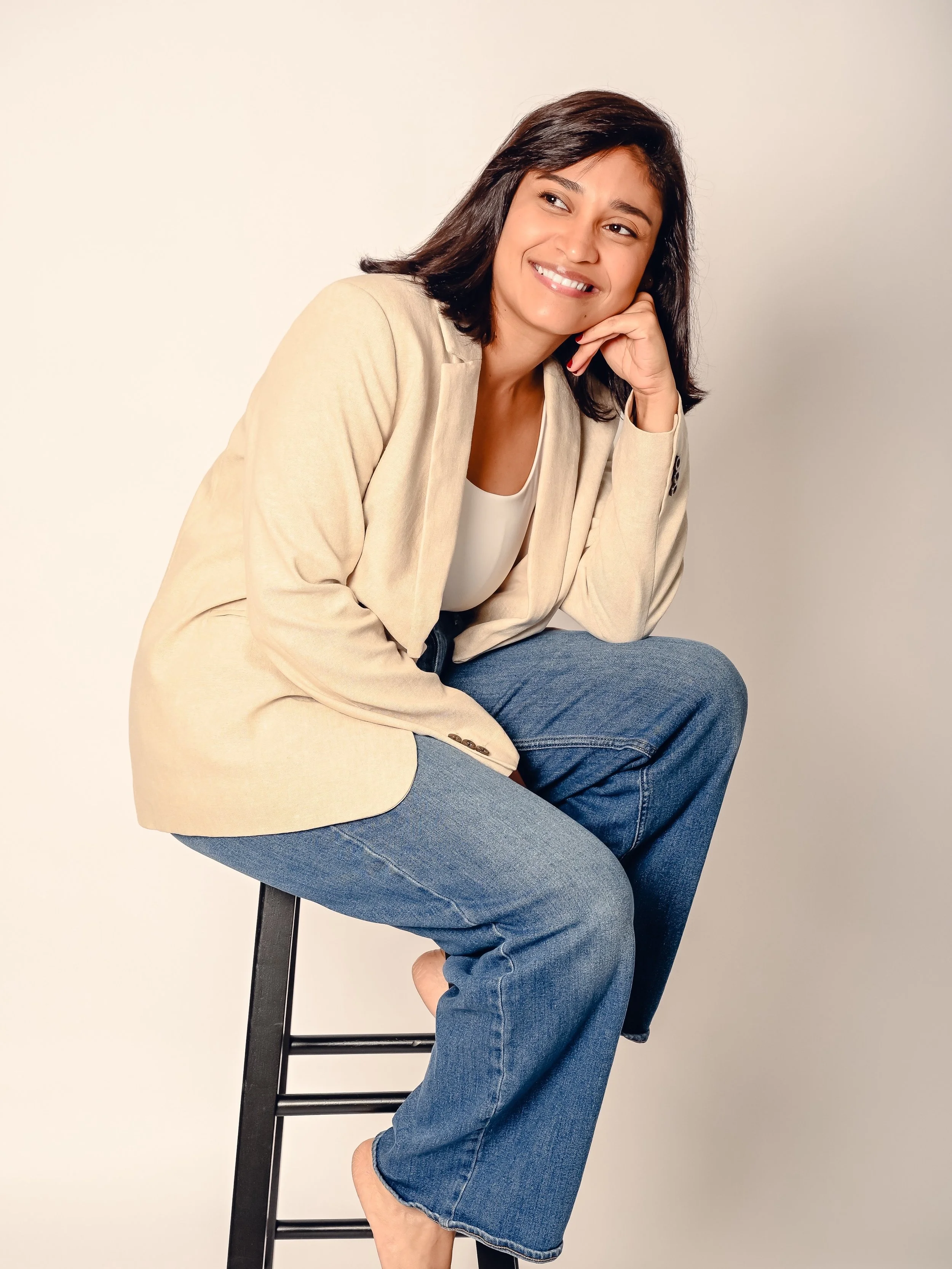 Mujer con cabello castaño oscuro y sonrisa, sentada en una silla negra, usando blusa blanca, chaqueta beige y jeans azules, en un fondo blanco.