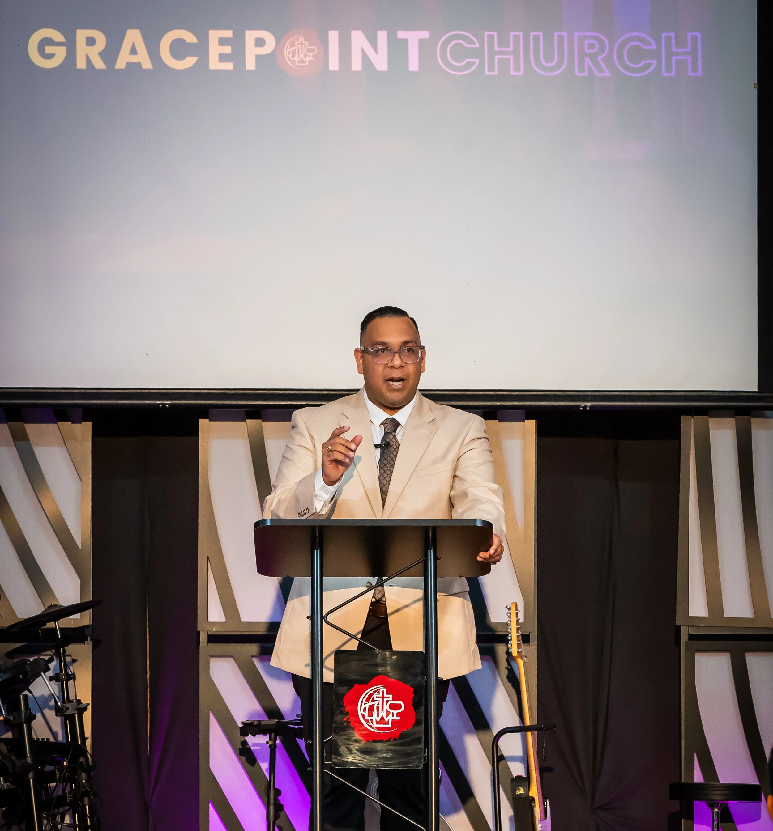 Un hombre con gafas y saco beige está dando un sermón en un púlpito y detrás hay una pantalla que dice 'GracePoint Church'.