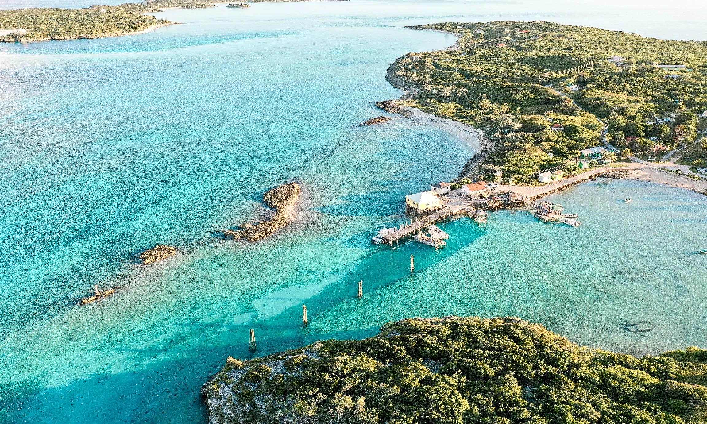 Aerial view of a tropical island coastline with turquoise water, rocky coves, sandy beaches, docks, boats, and lush vegetation.