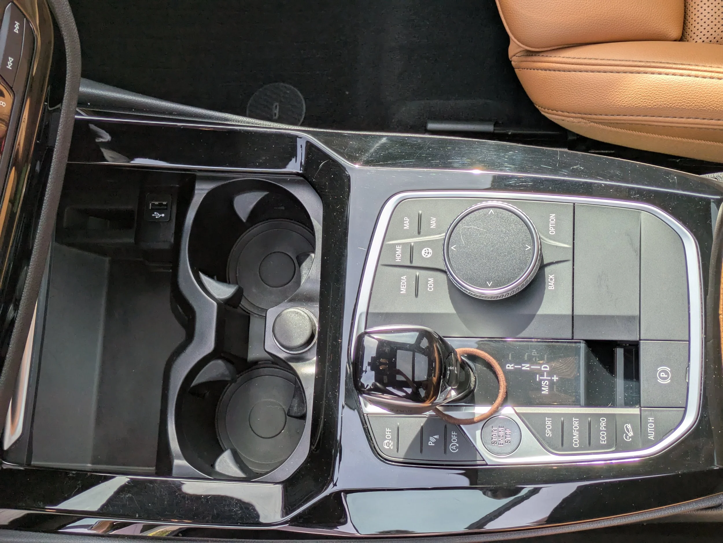 Center console of a BMW car showing a gear shifter, control buttons, and a small storage compartment, with a brown leather car seat visible in the background.