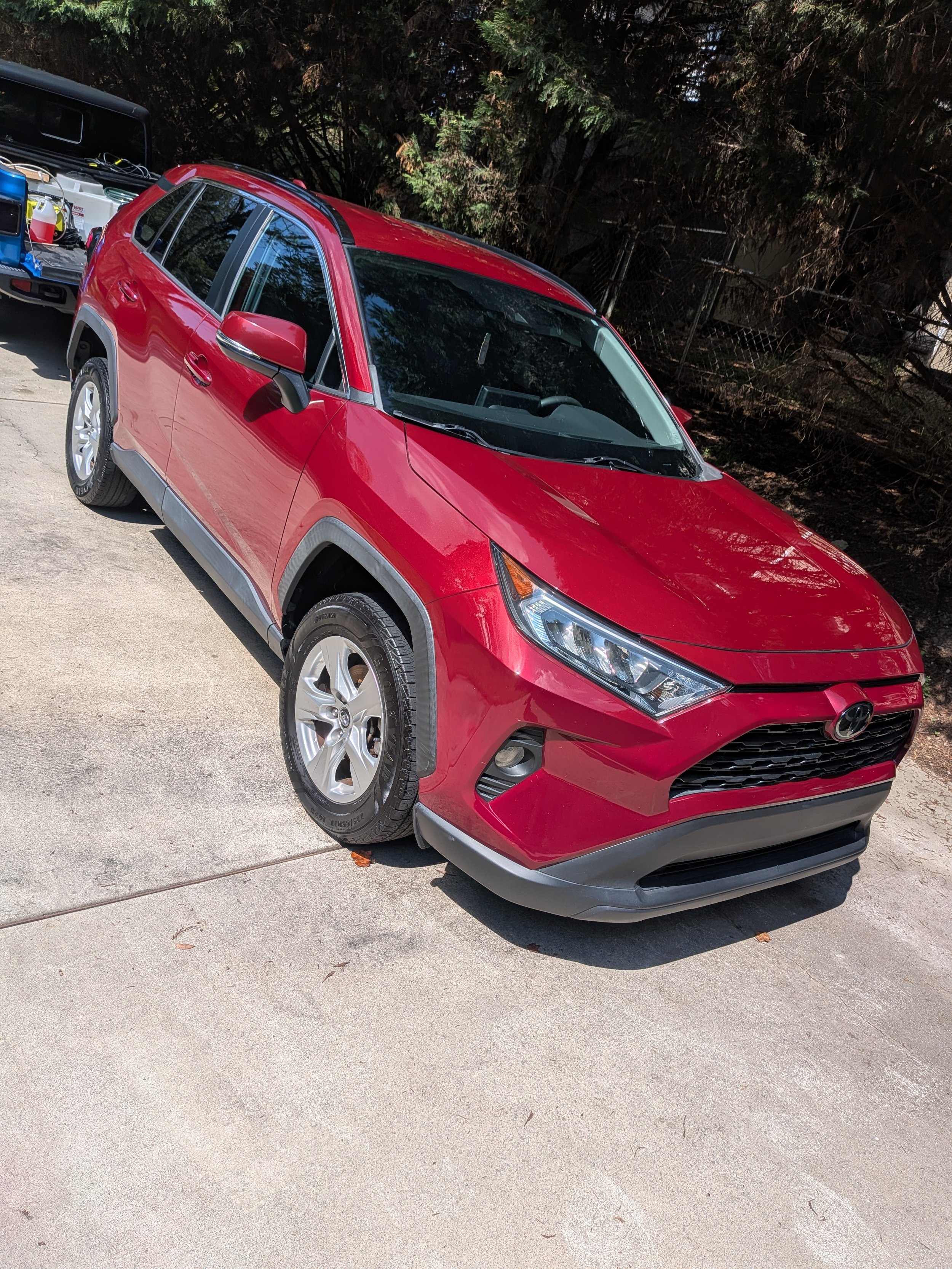 A red compact SUV parked on a concrete driveway with trees in the background and a small utility trailer attached, containing various items.