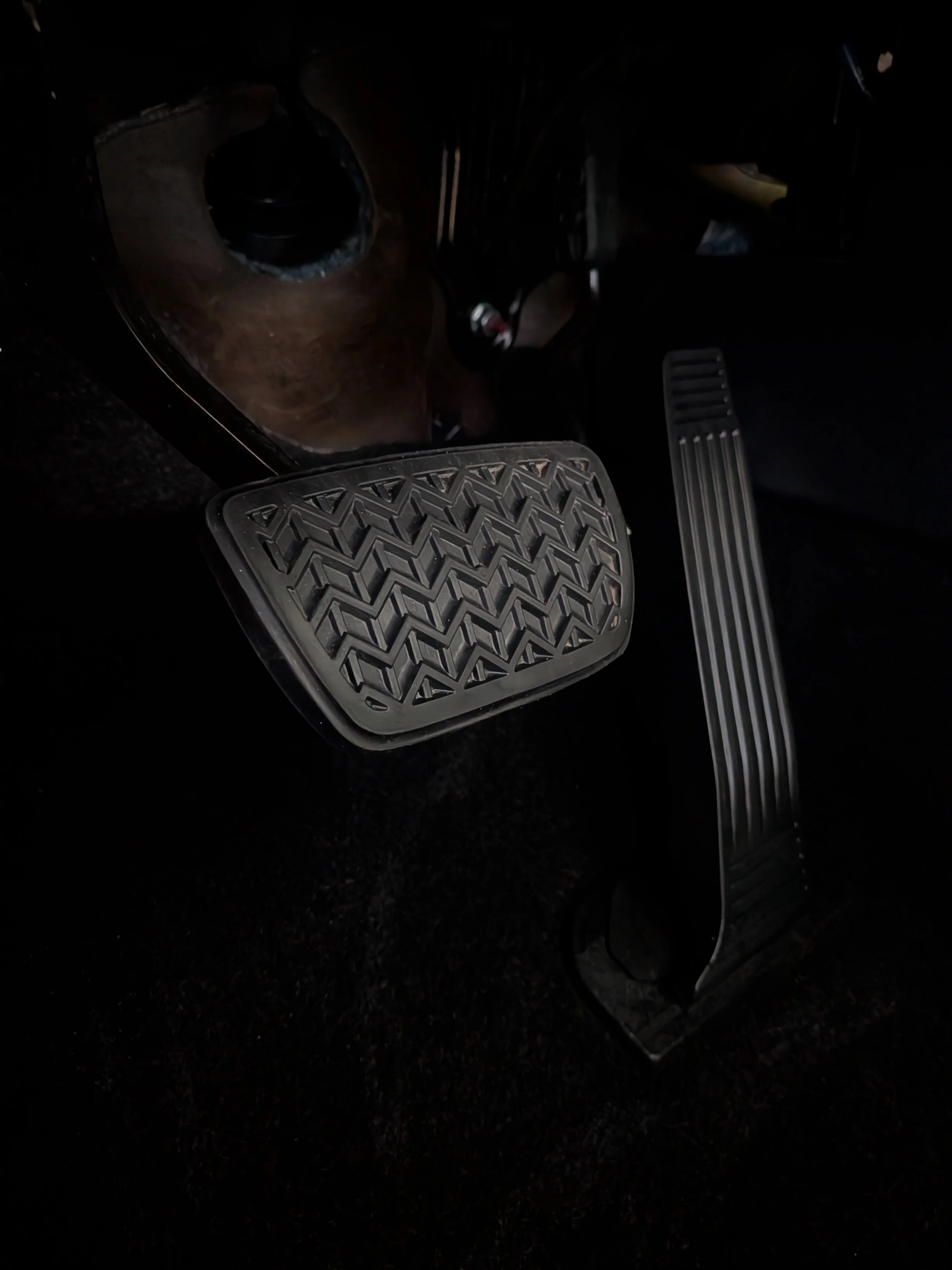 Interior of a car showing the brake and accelerator pedals, with a small wooden table or platform in front of them, placed on a black floor.