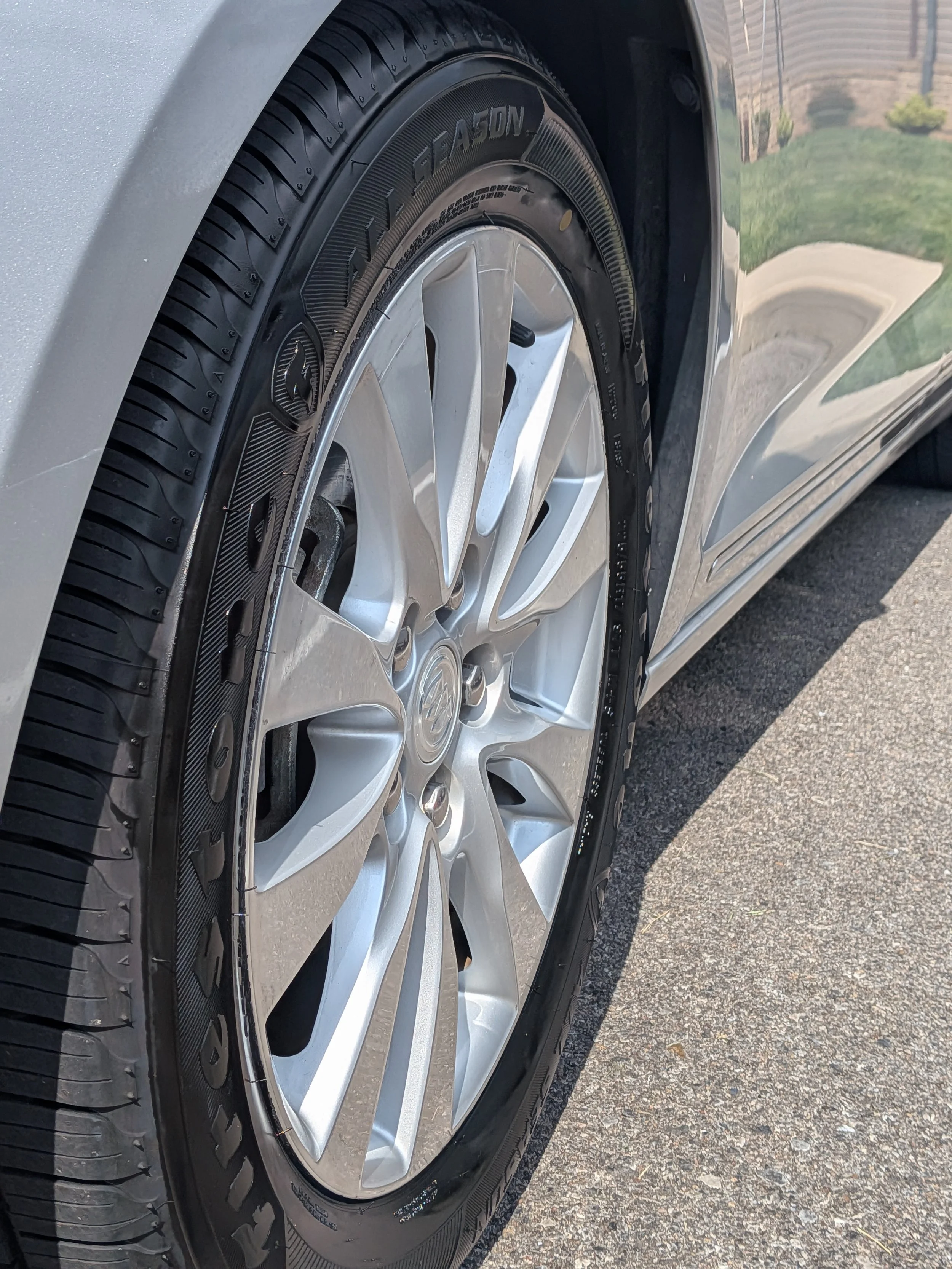 Close-up of a car tire and rim parked on a driveway with grass and a fence in the background.