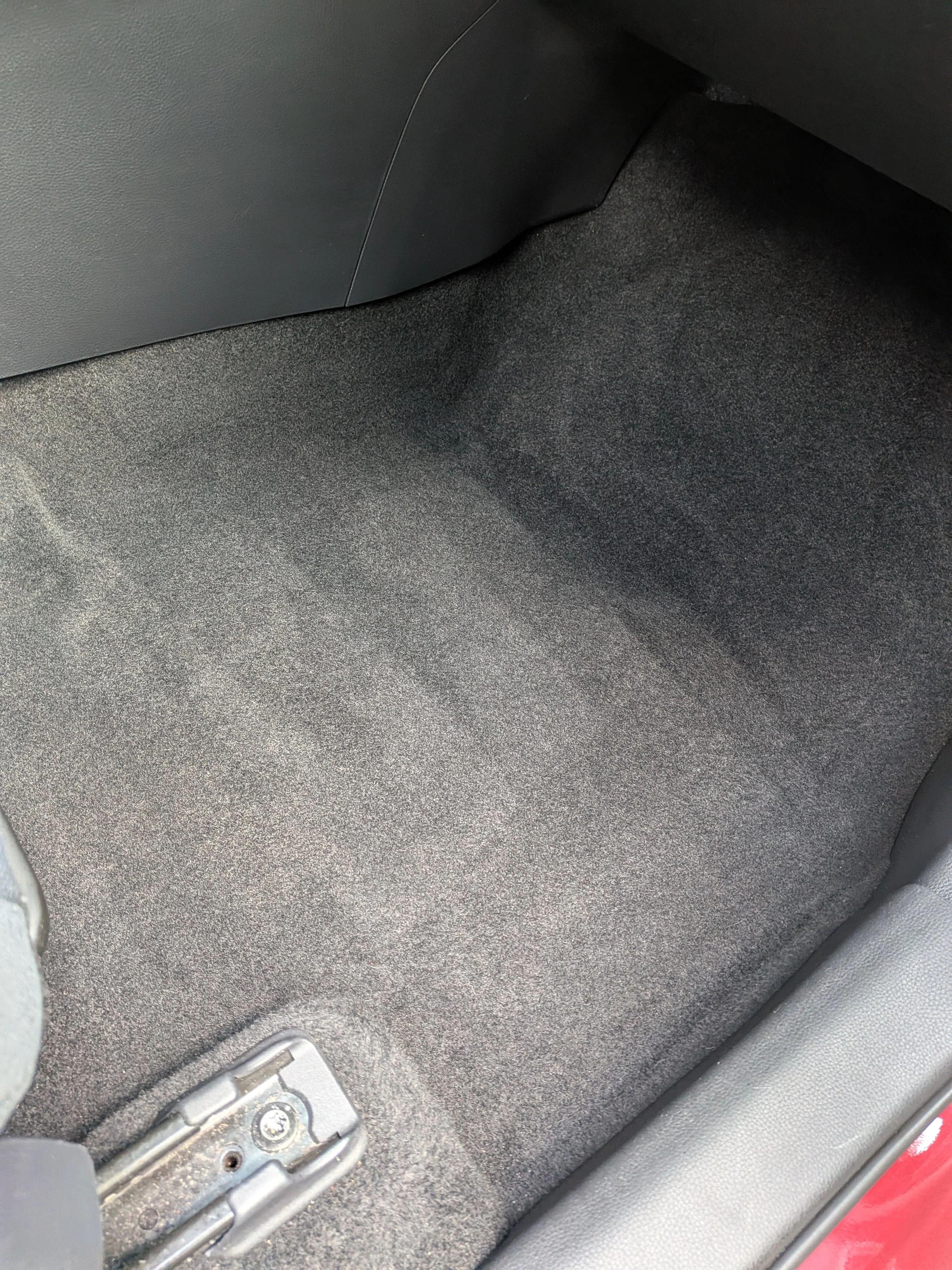 Close-up of car interior carpet with metal latch or hinge at the bottom, black textured flooring, and part of a red door visible at the bottom right.