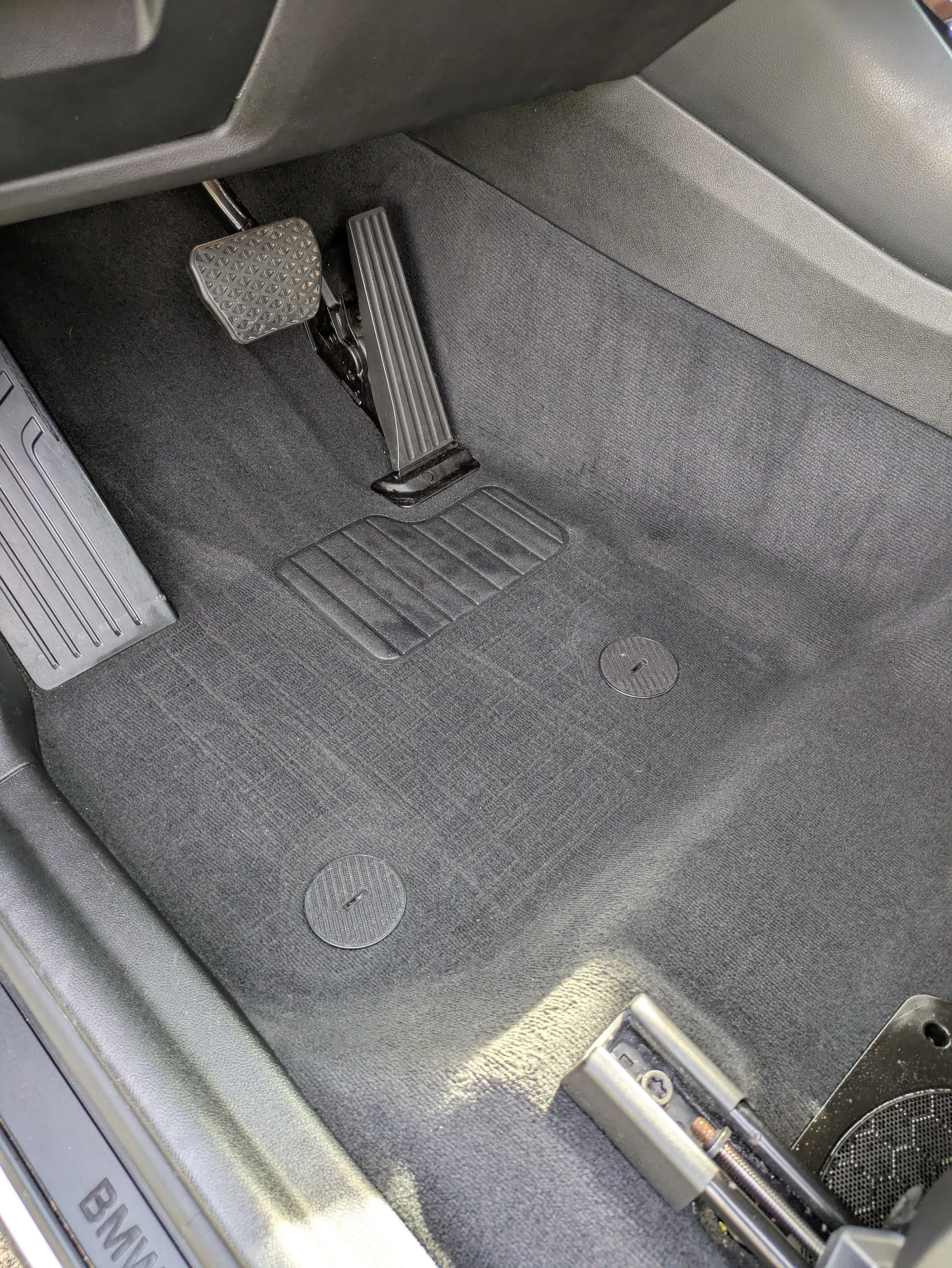 Close-up of the driver's side floor of a BMW vehicle showing the accelerator pedal, brake pedal, and footrest, all made of metal and rubber, with part of the car's interior and door visible.
