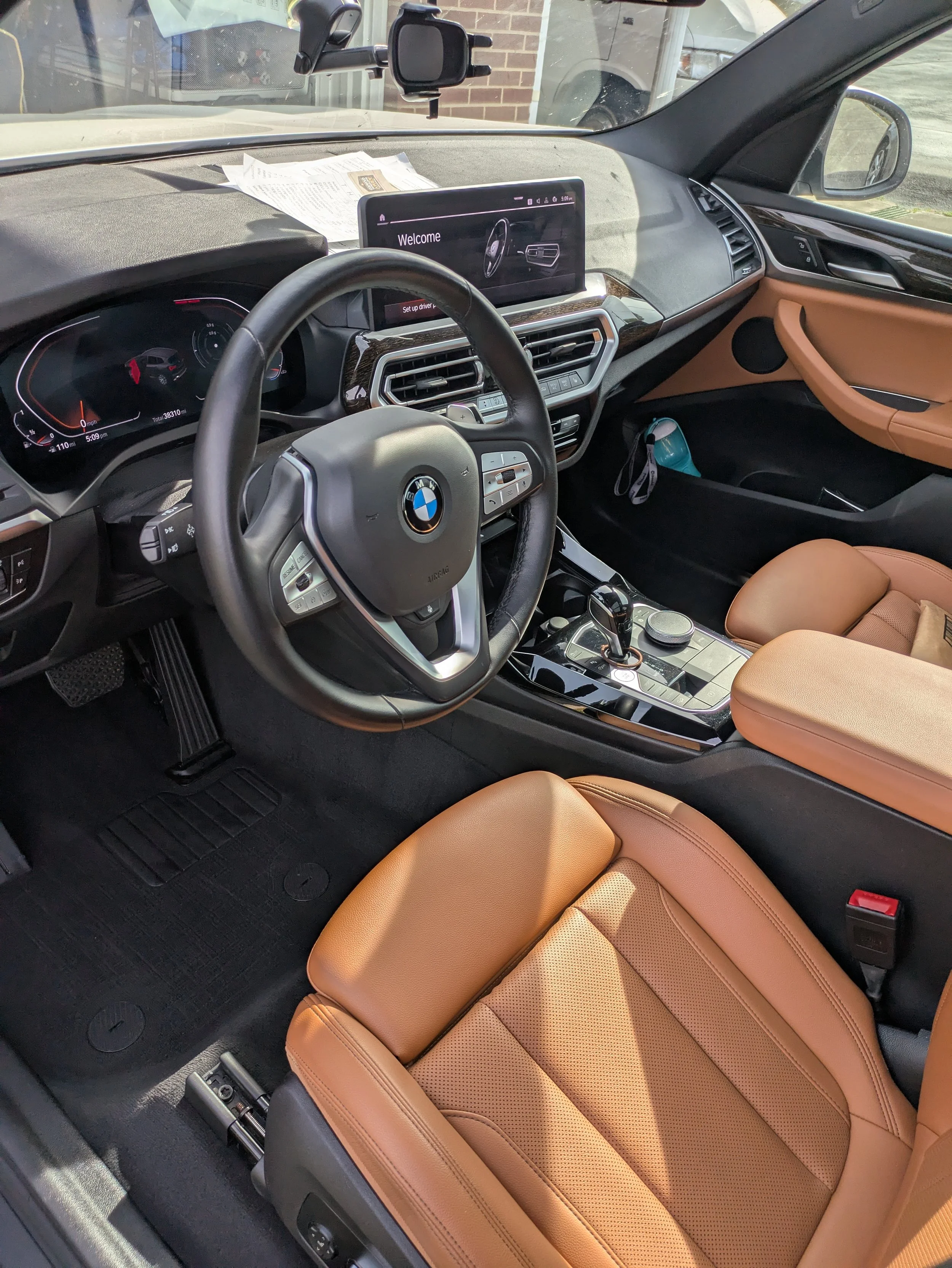 Interior of a BMW vehicle showing tan leather seats, a steering wheel with the BMW logo, a digital dashboard, and a large central screen displaying a welcome message. The dashboard and center console are black with metallic accents, and there is a paper on the dashboard.