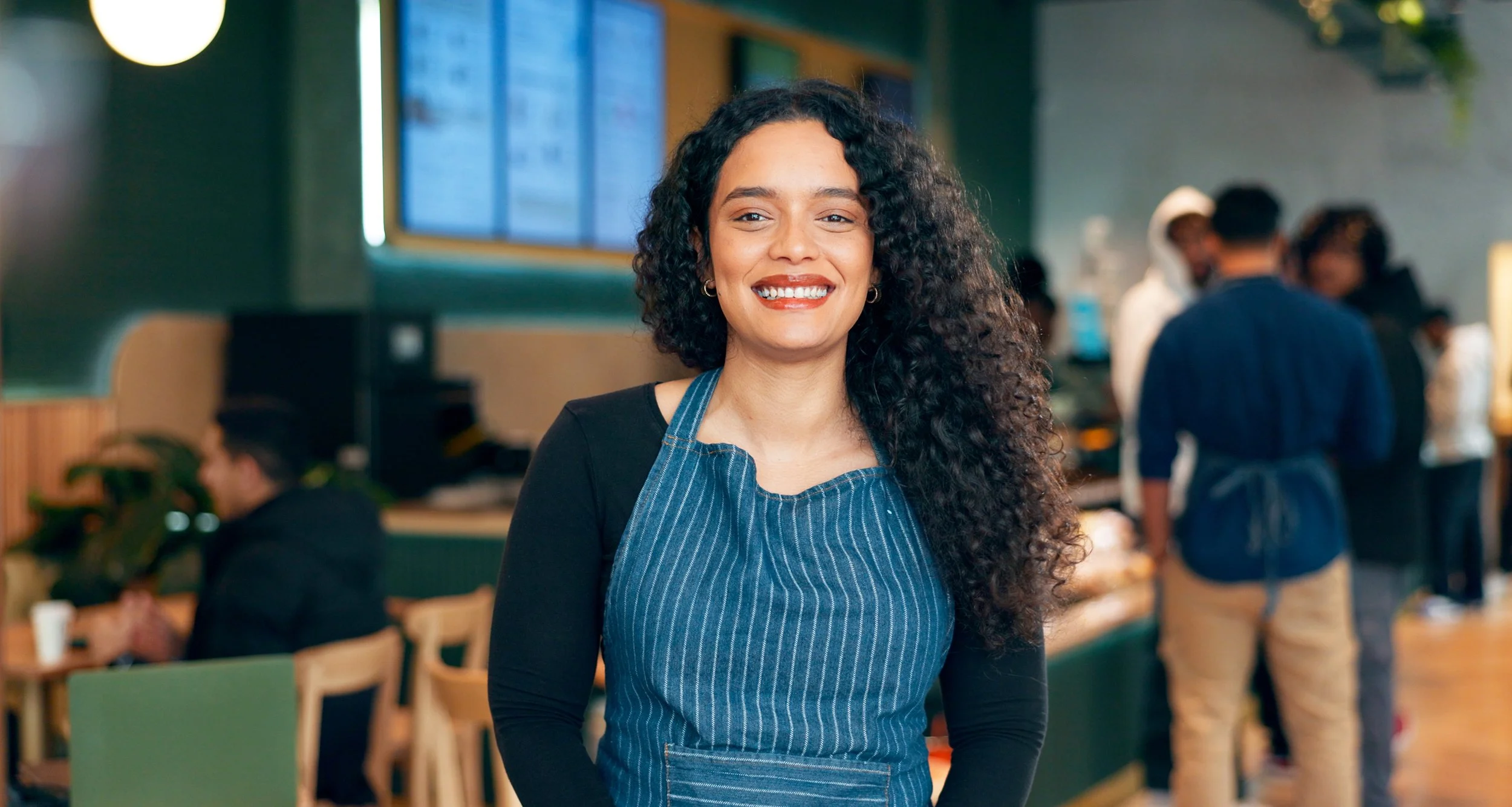 Learn It Media. A smiling woman with curly black hair wearing a blue apron in a busy restaurant or cafe setting.