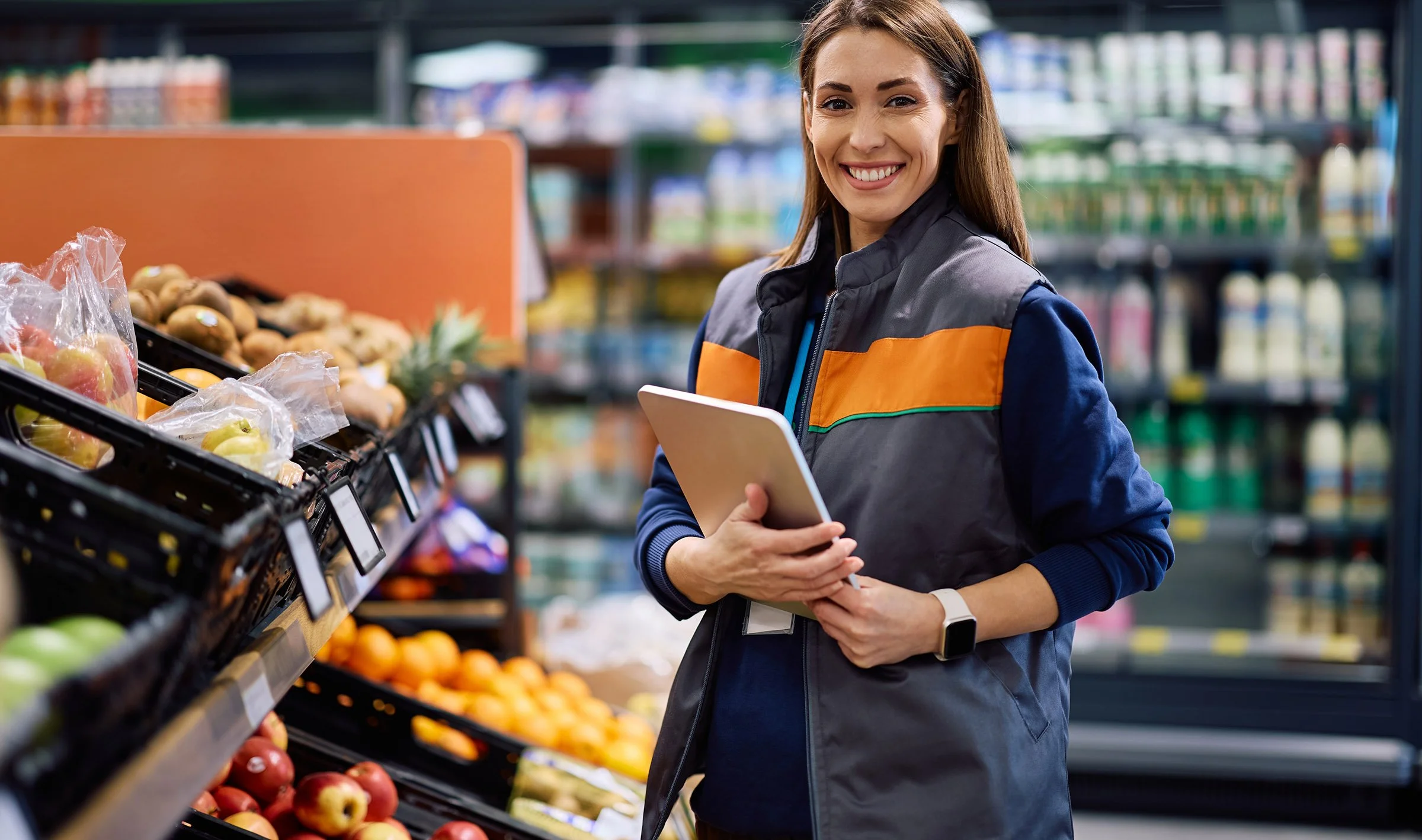 Learn It Media. A woman shopping for apples in a grocery store, holding a tablet, and smiling at the camera.