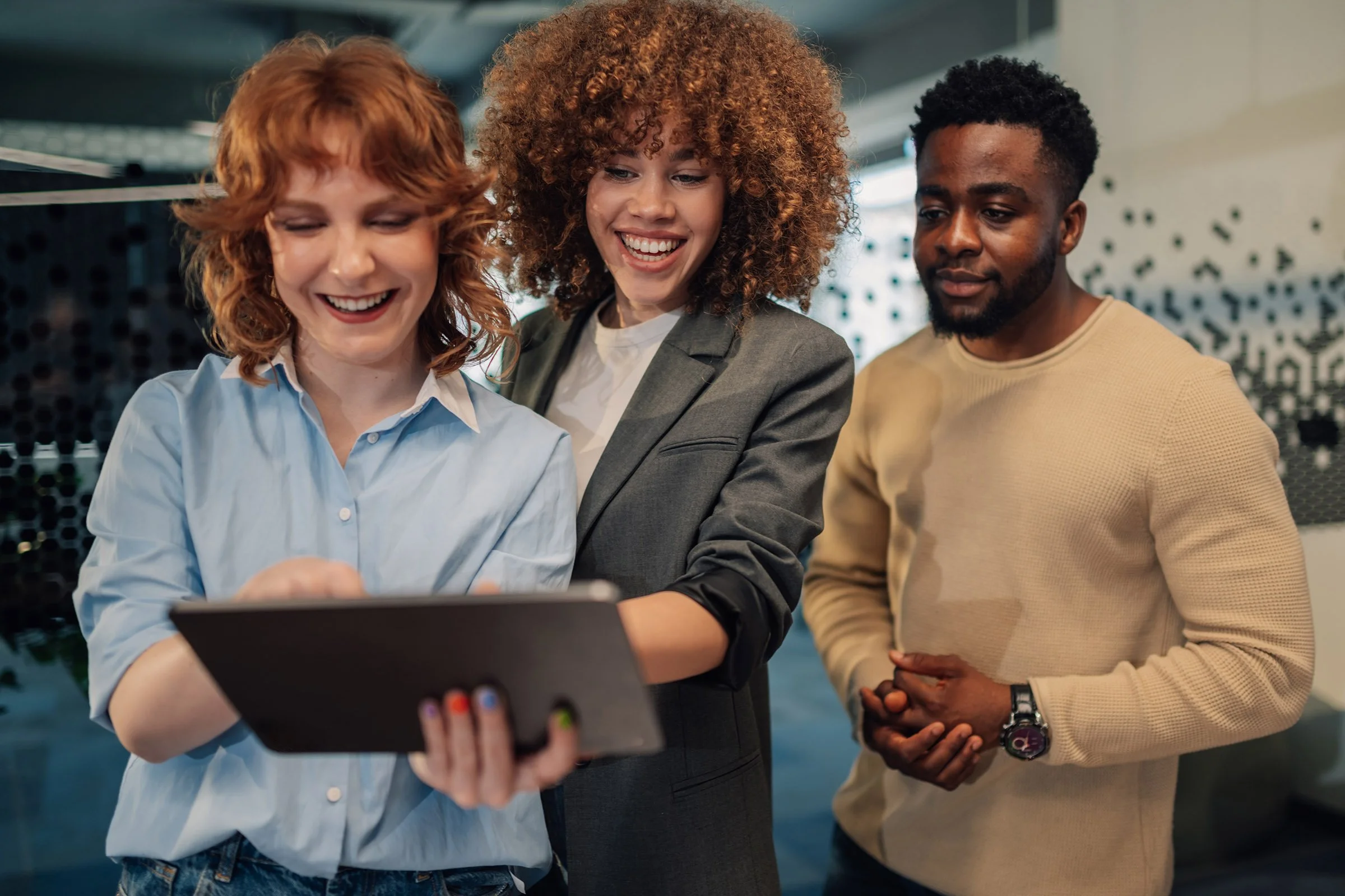 Learn It Media. Three diverse young adults looking at a tablet together, smiling, in a modern indoor setting.