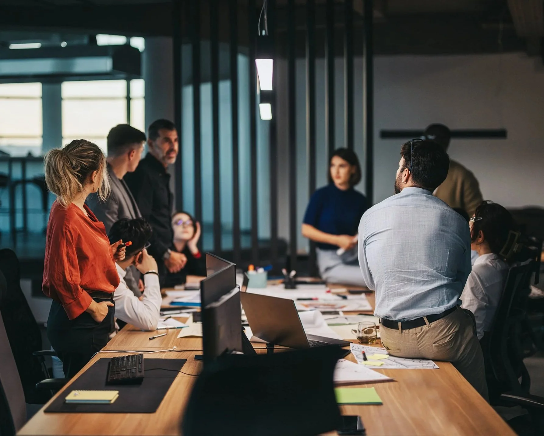 Learn It Media. A diverse group of professionals in a business meeting or presentation in a modern office, engaging in discussion around a large table with laptops, notebooks, and documents.