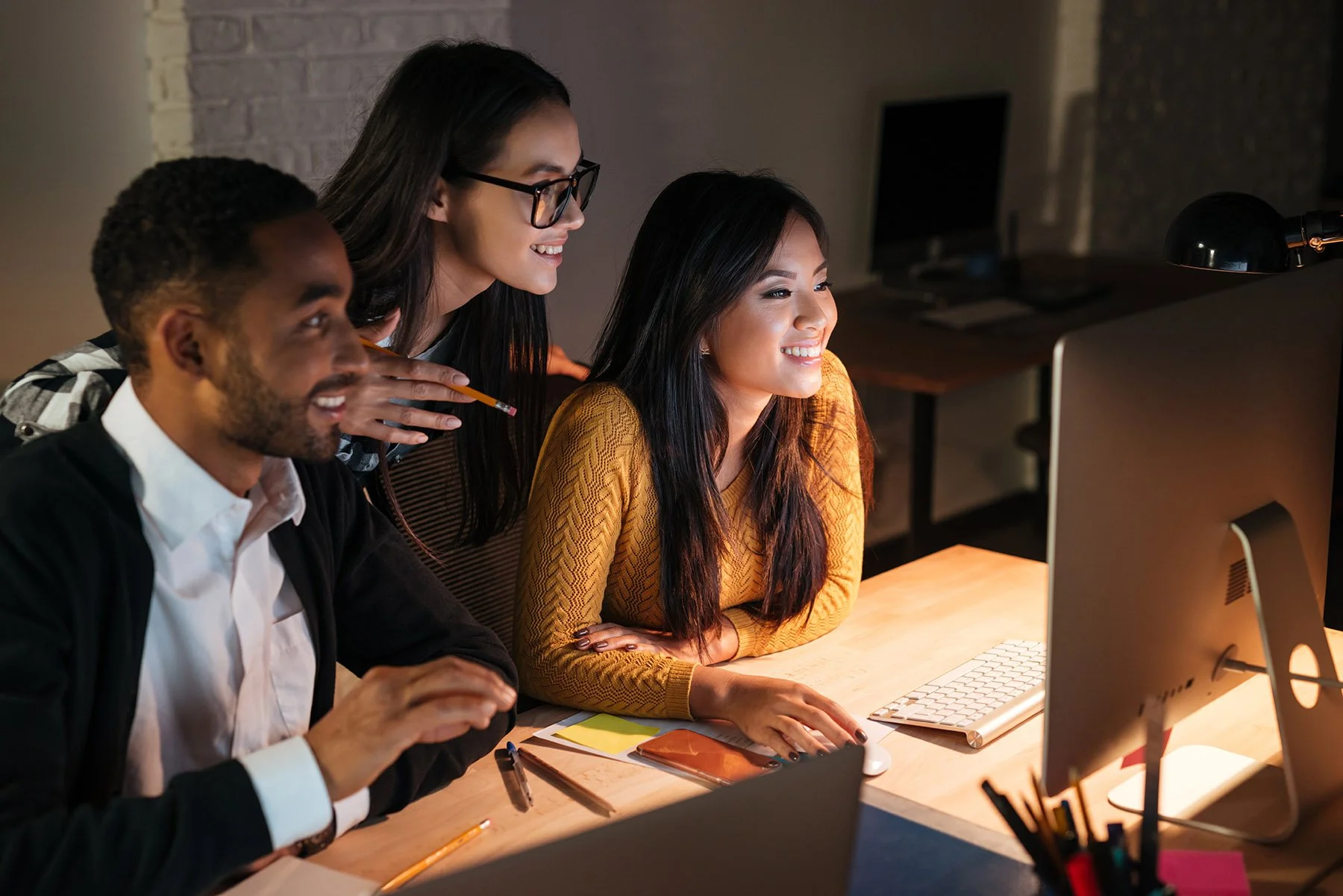 Learn It Media. Three people gathered around a computer screen in a dimly lit office, smiling and collaborating.