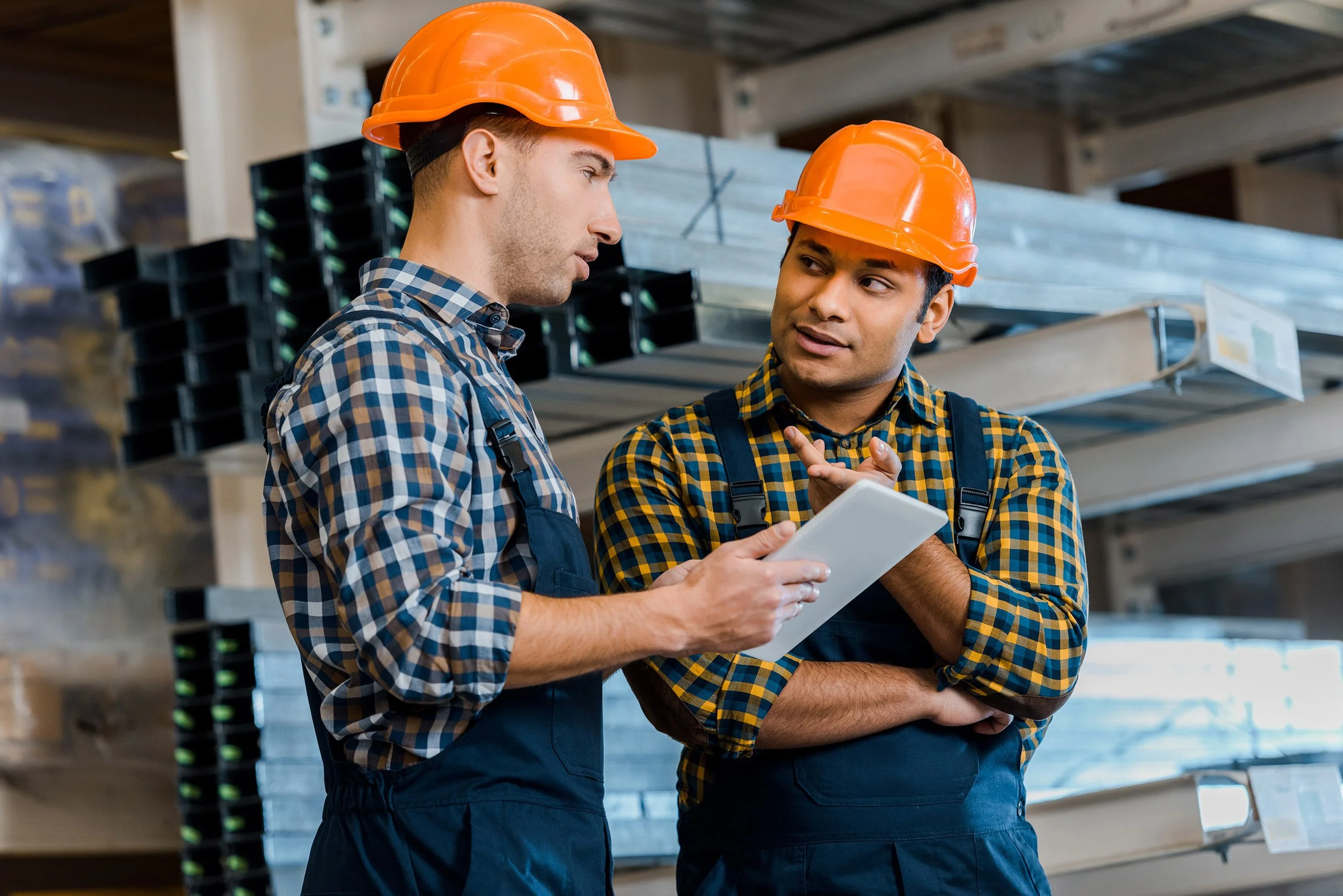 Learn It Media. Two construction workers wearing orange safety helmets and plaid shirts are discussing something while standing in a storage area with wood and metal racks.