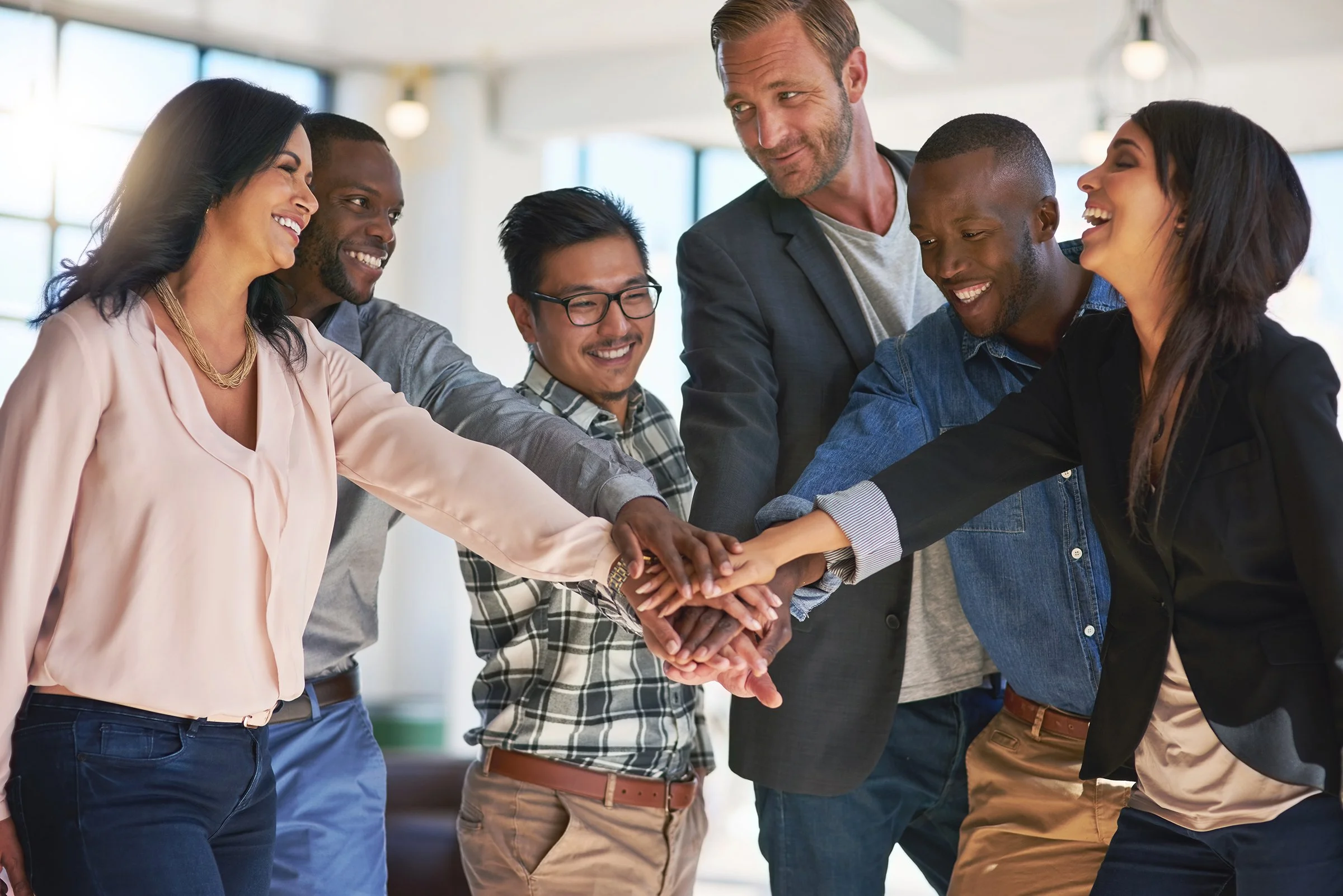 Learn It Media. Group of diverse people in business casual attire joining hands in a team-building gesture, smiling and showing camaraderie in an office setting.