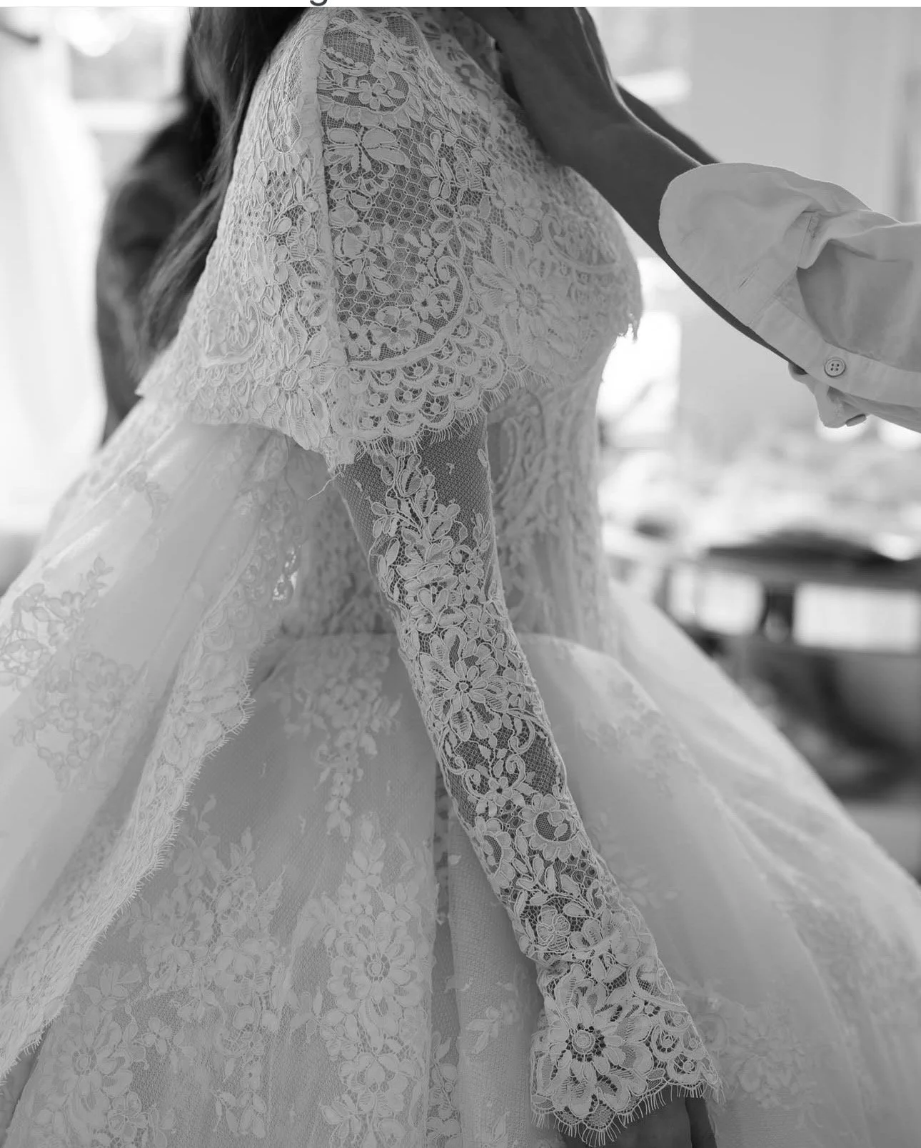 Donna in abito da sposa ricamato con pizzo durante la preparazione, con un'altra persona che la aiuta.