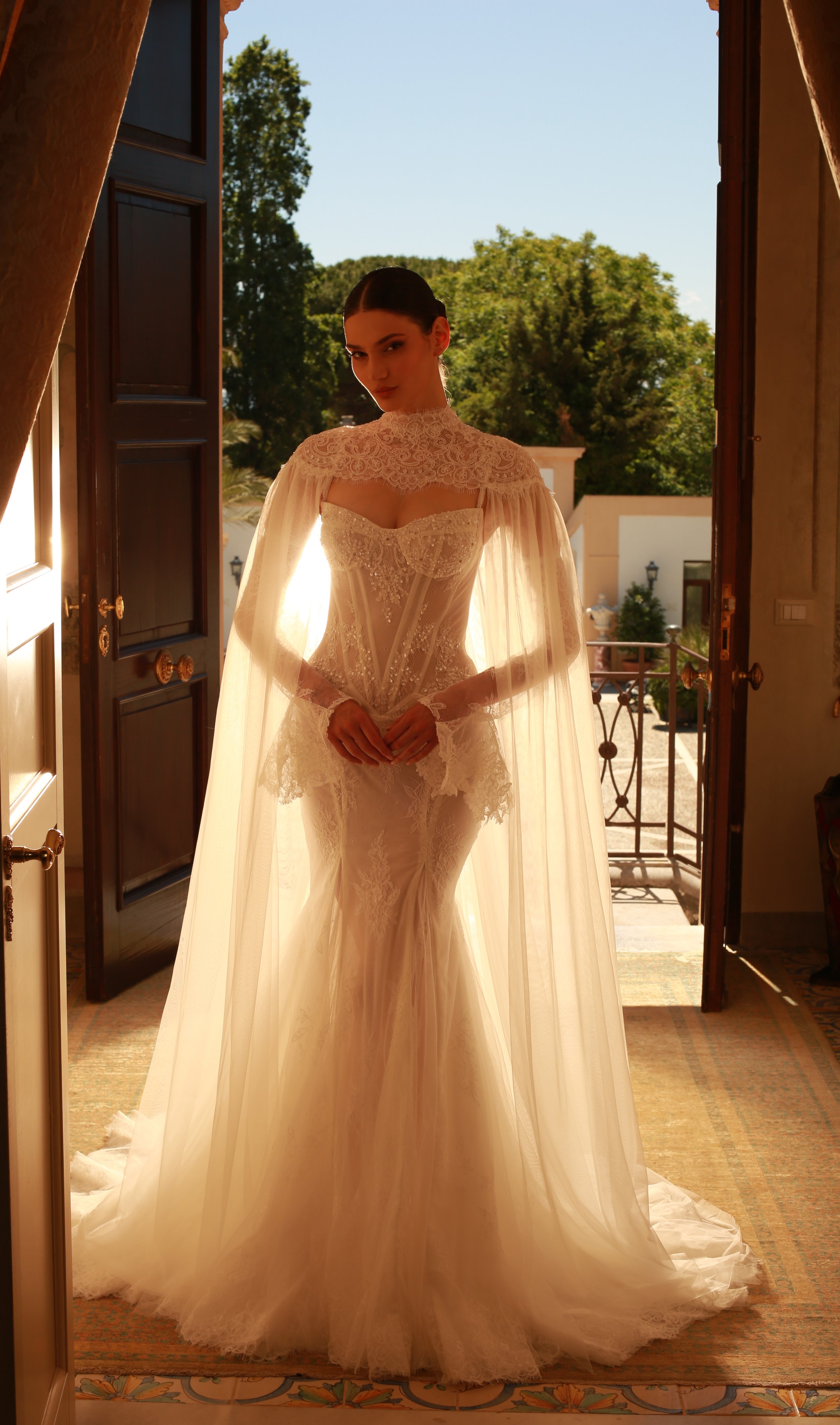 Una donna in abito da sposa bianco con dettagli in pizzo, che si staglia di fronte a una porta aperta che dà su un balcone con un cielo blu e alberi verdi sullo sfondo.