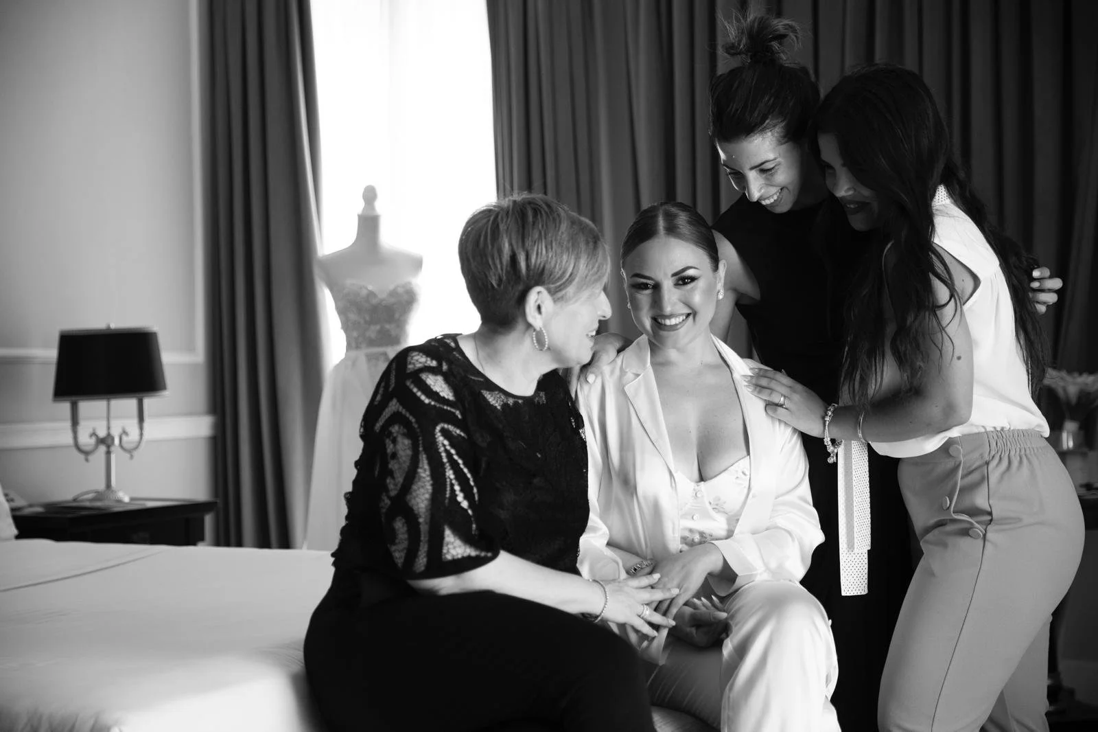Cinque donne, di cui una con abito bianco, sorridono e si abbracciano in una stanza elegante con venerdì in soffitto e tende. Una delle donne con abito bianco sembra essere la sposa, circondata da amici e parenti che la aiutano e si congratulano con lei.