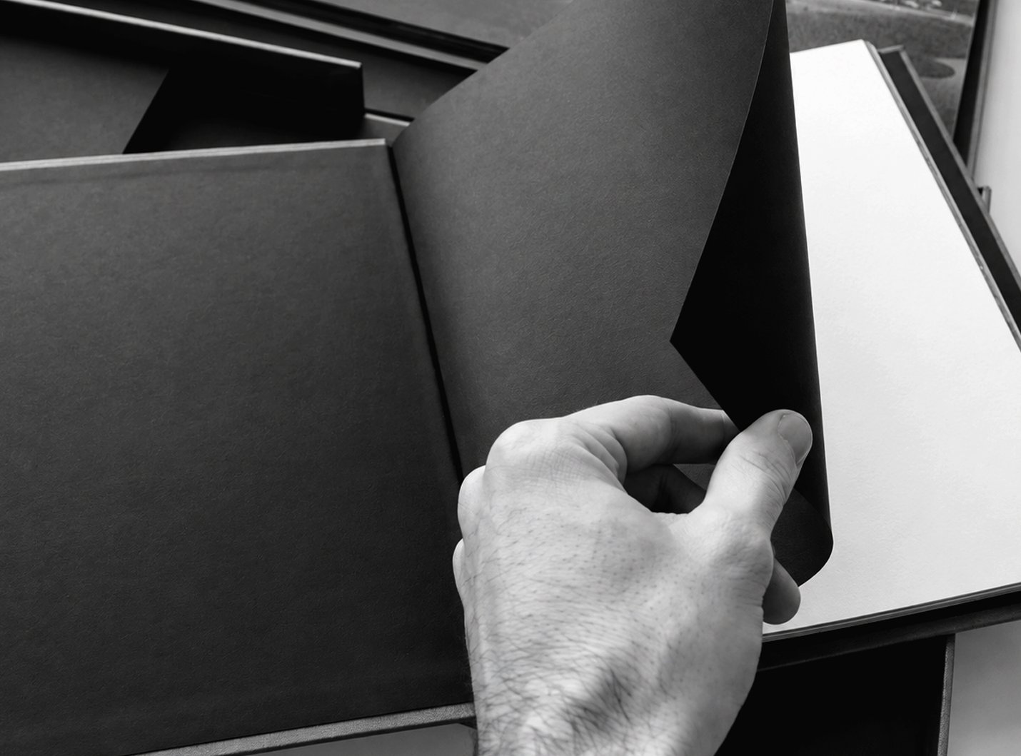 Pablo's hand flipping through the pages of a black hardcover book, with a blank white page visible on the right side, on a desk near a window.
