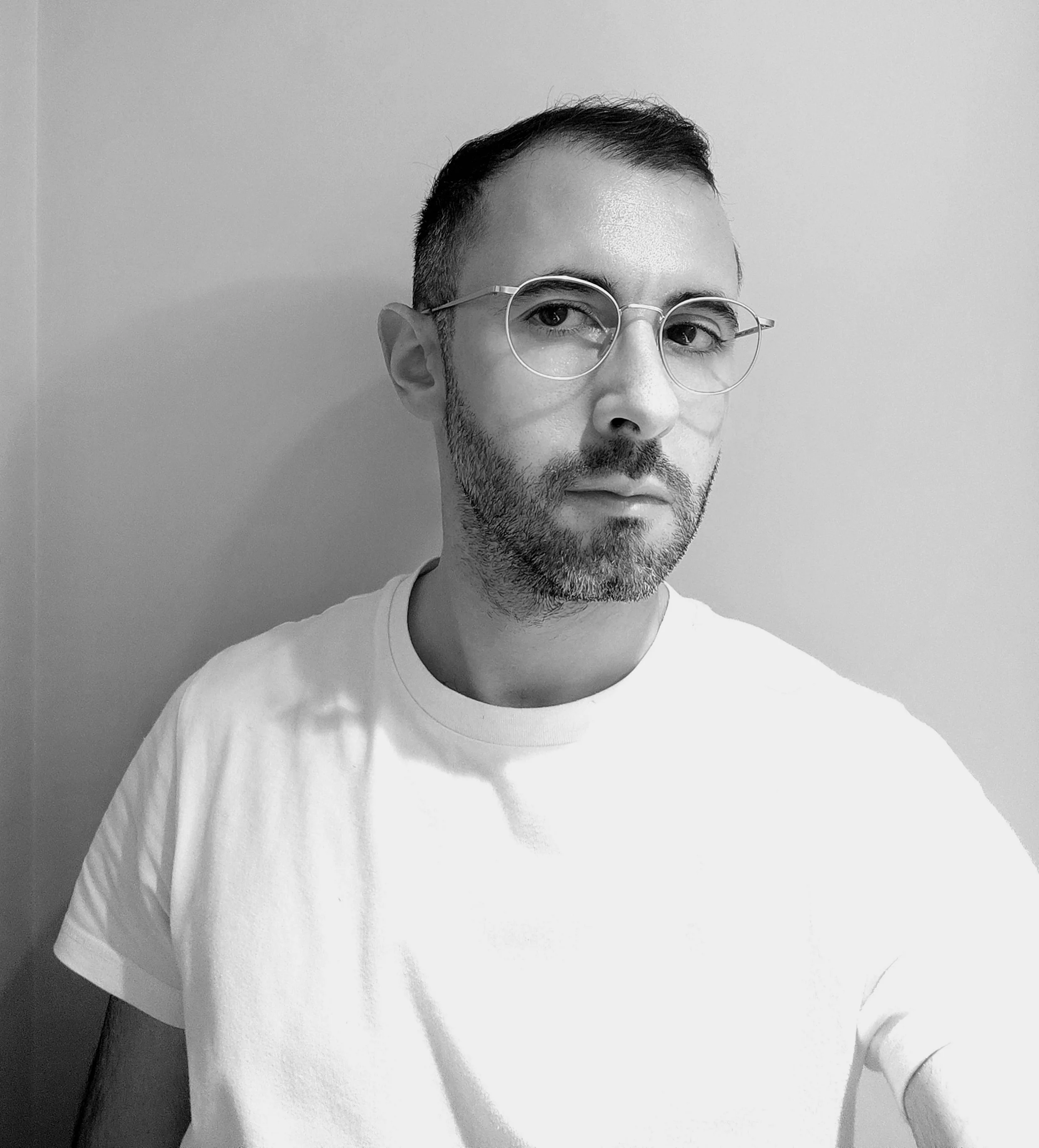Black-and-white photo of Pablo with glasses, short hair, and a beard wearing a white t-shirt, standing against a plain wall.