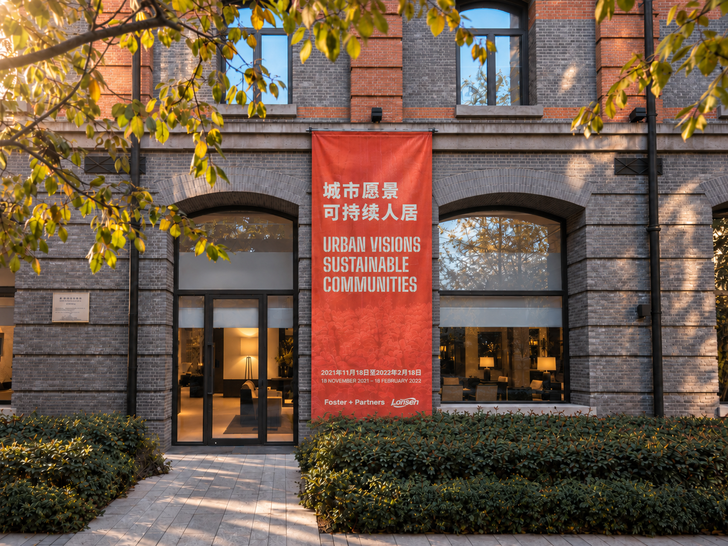 Outdoor orange banner showing the exhibitiopn branding in dual language. The vertical banner hangs from the first floor.