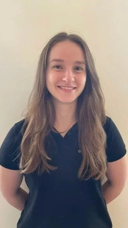A young woman with long light brown hair, smiling, wearing a black polo shirt, standing against a plain off-white wall.