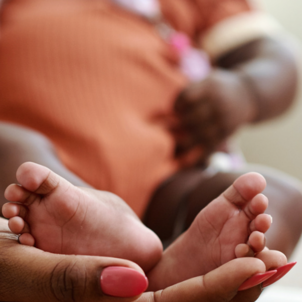Close-up of a newborn baby's feet held gently by an adult's hand with pink-painted nails, blurred background showing a person holding the baby.