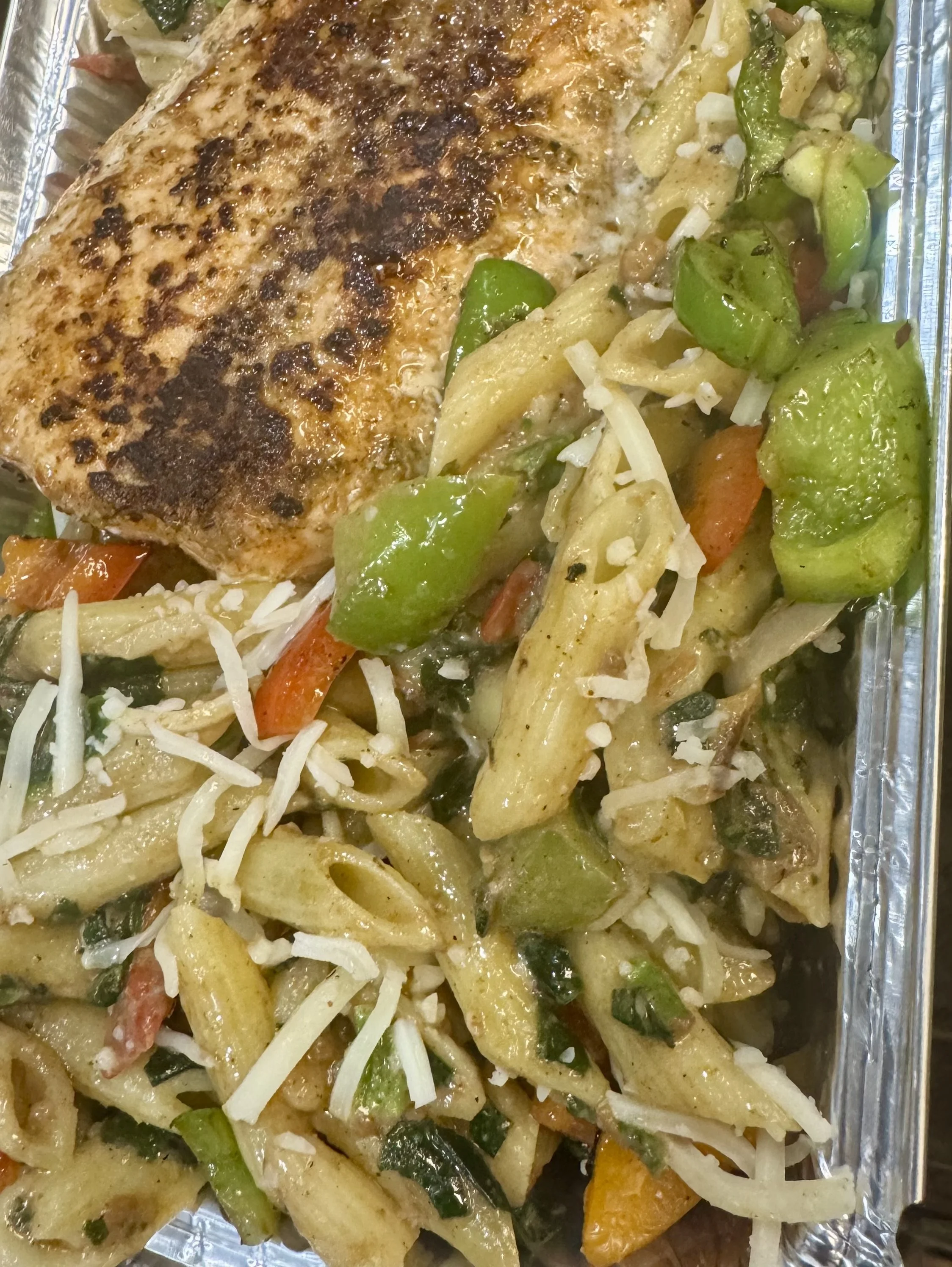 Close-up of baked chicken breast with grilled surface, served with mixed roasted vegetables including bell peppers, zucchini, and carrots, in foil tray.