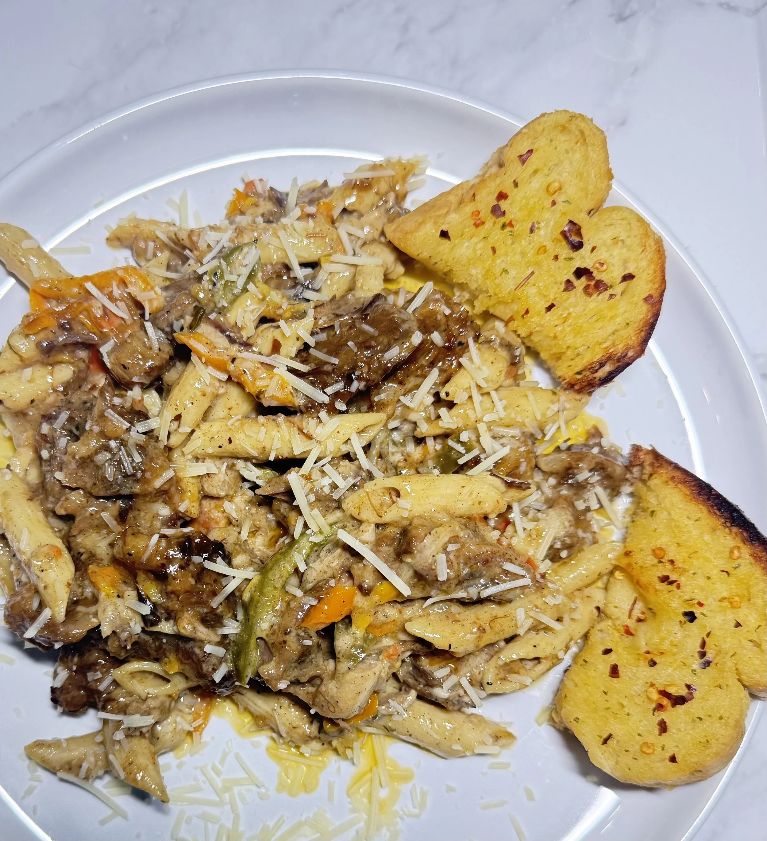 Plate of cooked pasta with a creamy mushroom and vegetable sauce, topped with shredded cheese, and two slices of garlic bread sprinkled with red pepper flakes.