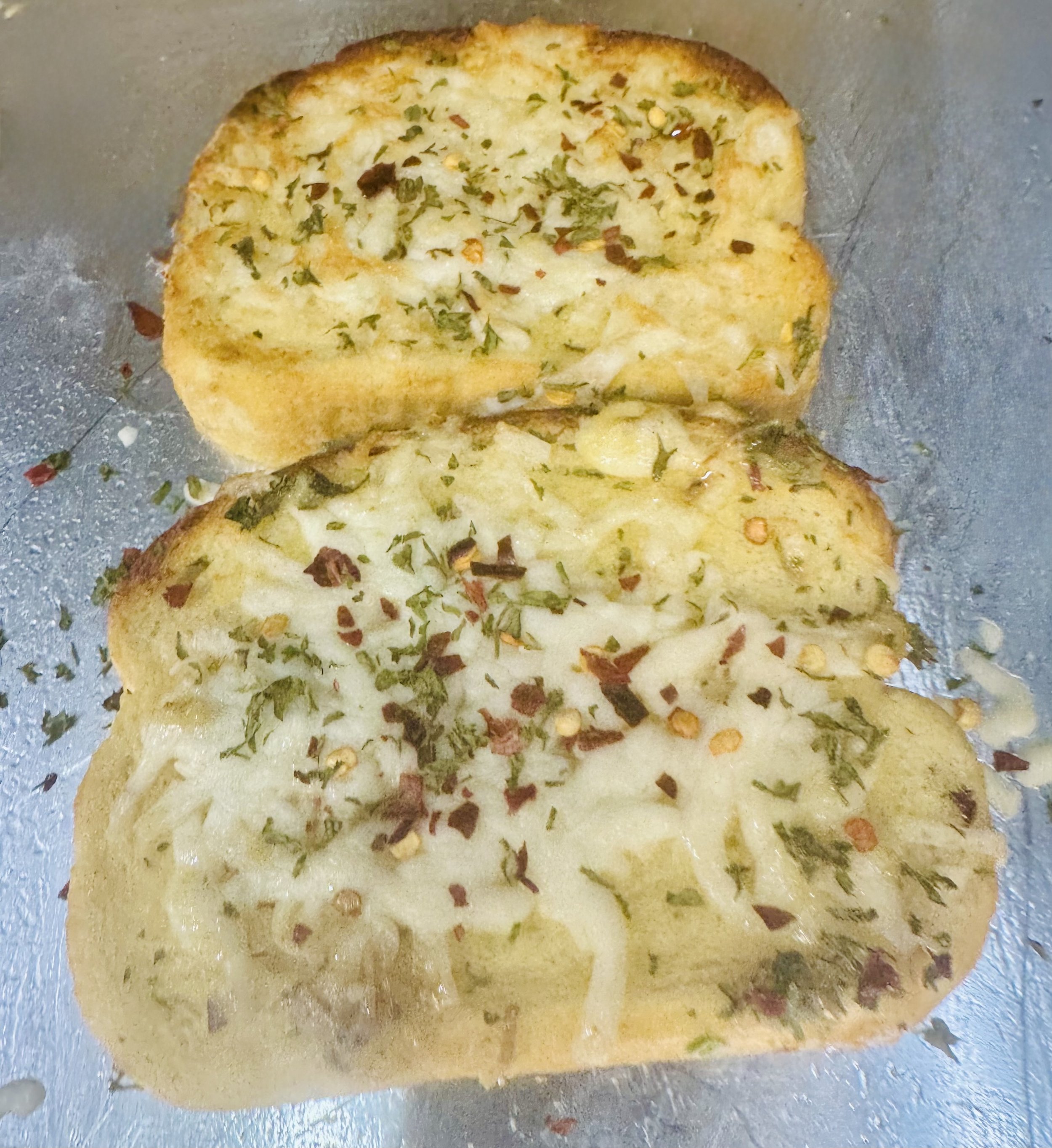 Two slices of garlic bread topped with parsley, cheese, and herbs on a baking sheet.