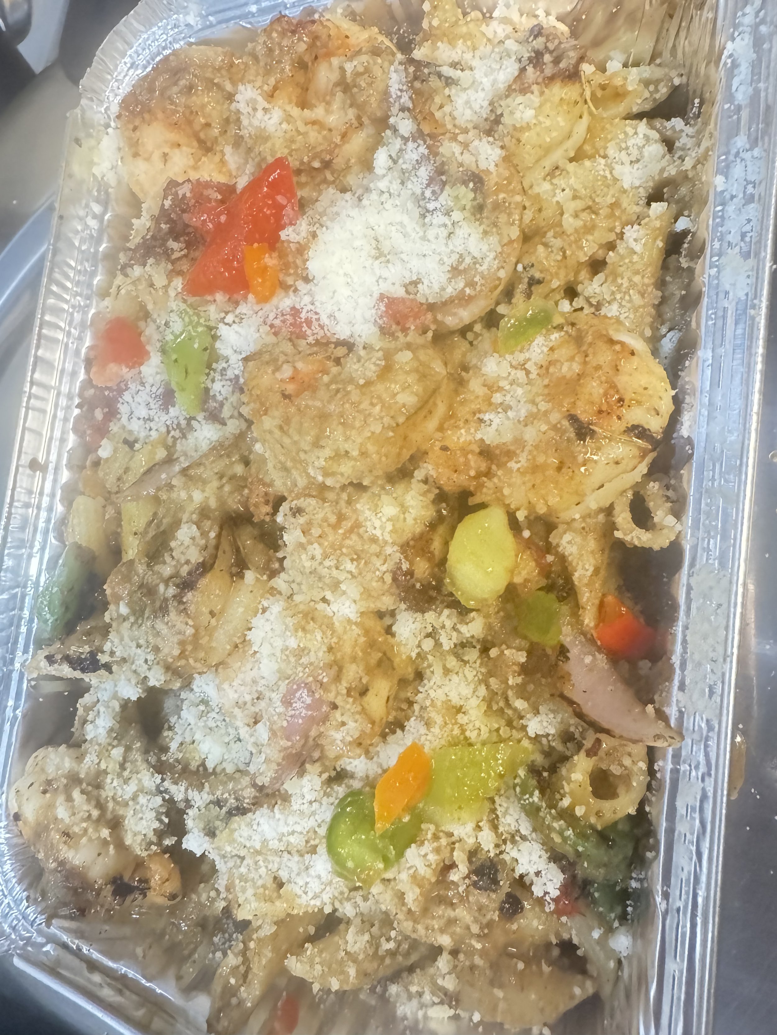 A disposable aluminum tray lined with clear plastic wrap containing cooked chicken pieces topped with riced cauliflower and garnished with diced carrots and celery.