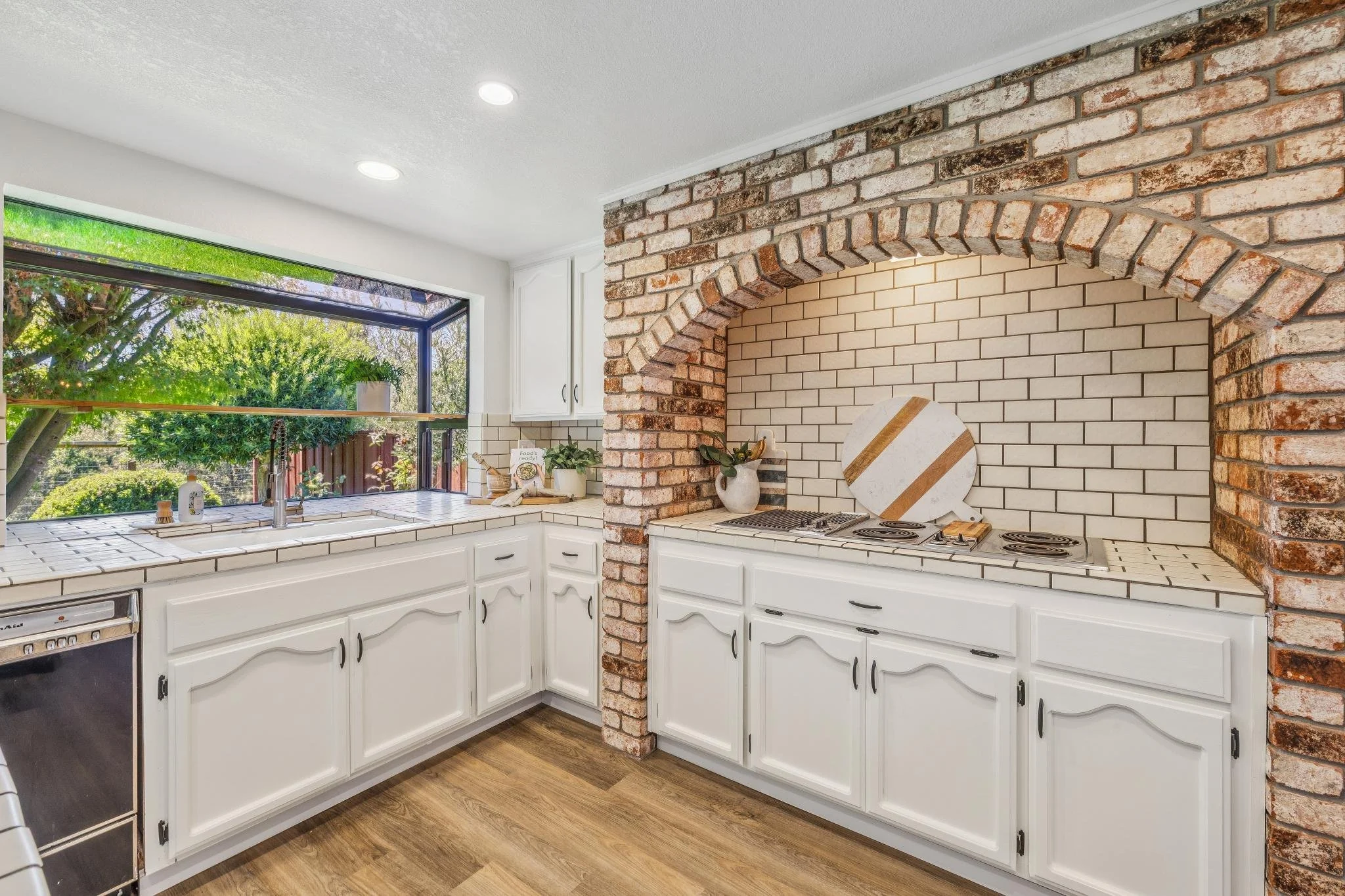 A kitchen with white cabinets, a white tiled countertop, and a brick accent wall with an arched brick opening. Large window with green trees outside, sunlight coming in.