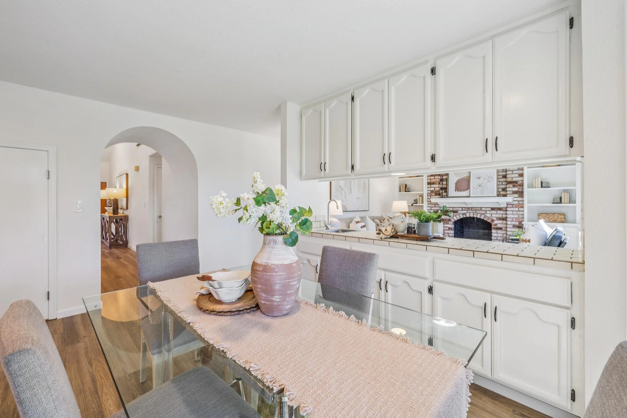 A dining area with a glass top table, gray upholstered chairs, a pink vase with white flowers, and a kitchen with white cabinets and a brick fireplace in the background.
