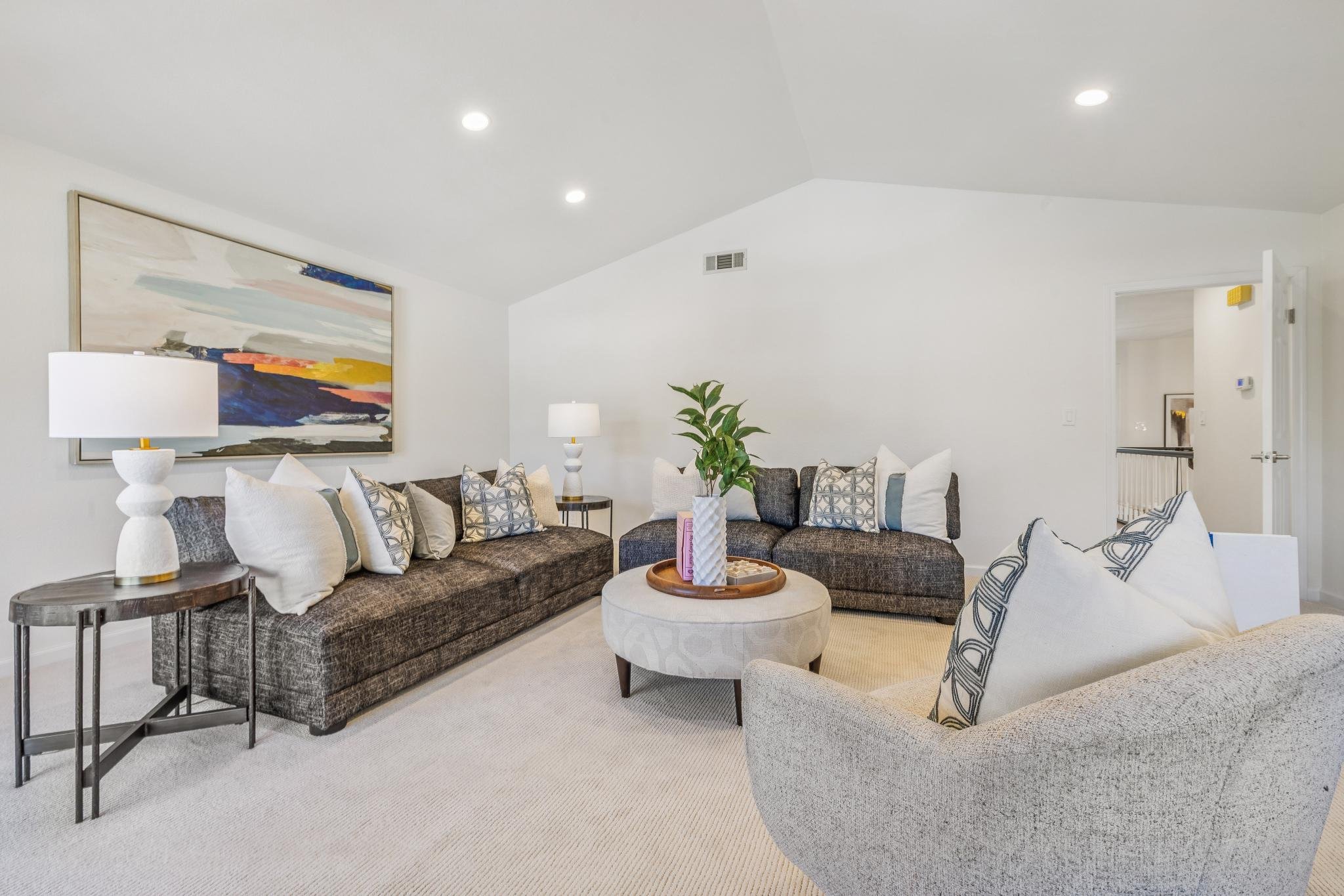 Living room with gray sofa, white pillows, black side tables, white table lamps, a white armchair, a round ottoman with a plant and books on it, abstract wall art, and white walls with recessed lighting.