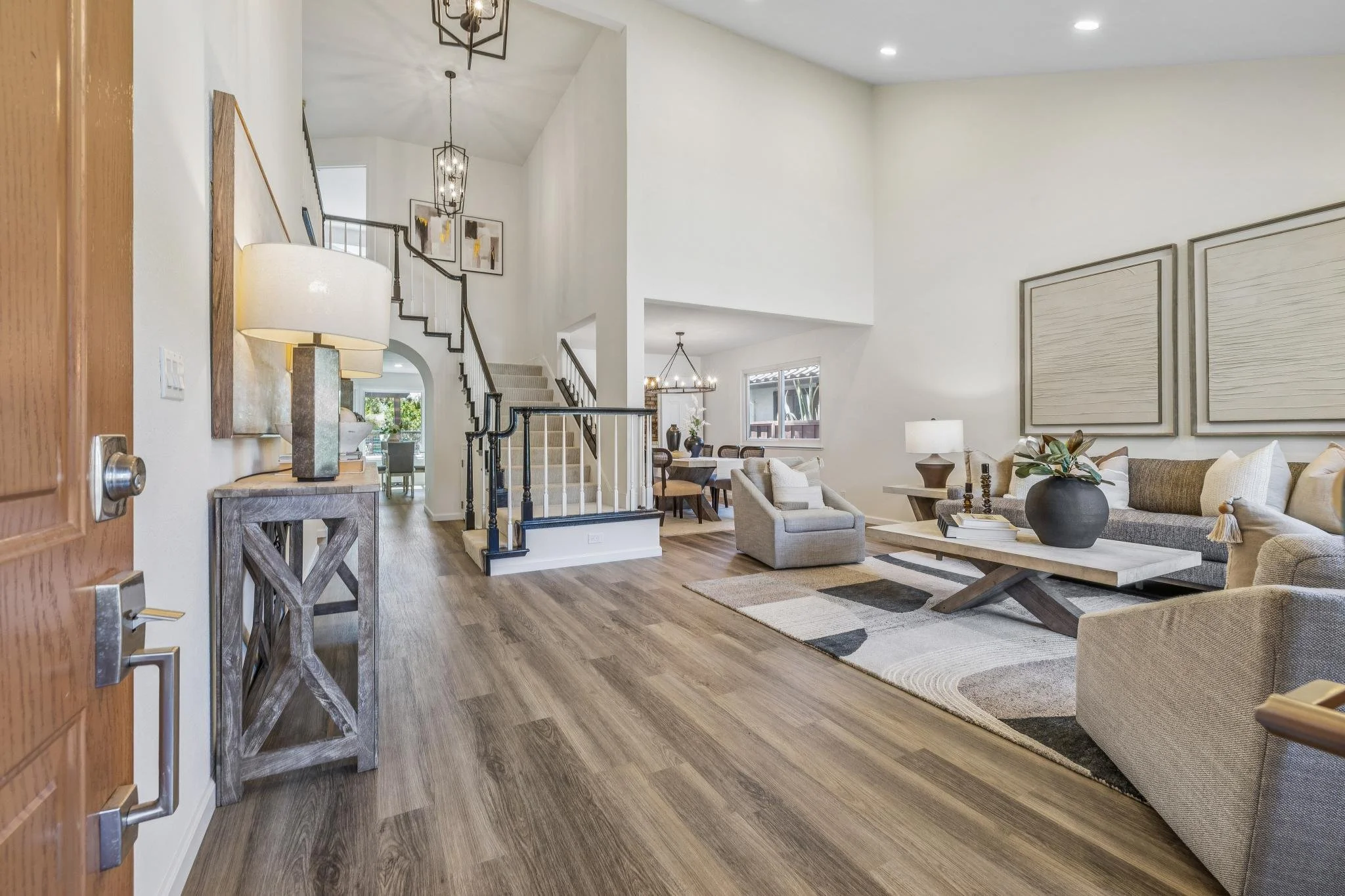 Living room with beige sofas, wooden coffee table, decorative vases, large framed artwork, and a rug. An open staircase with black railing leads to an upper level, and there's a dining area with a chandelier and a window in the background.