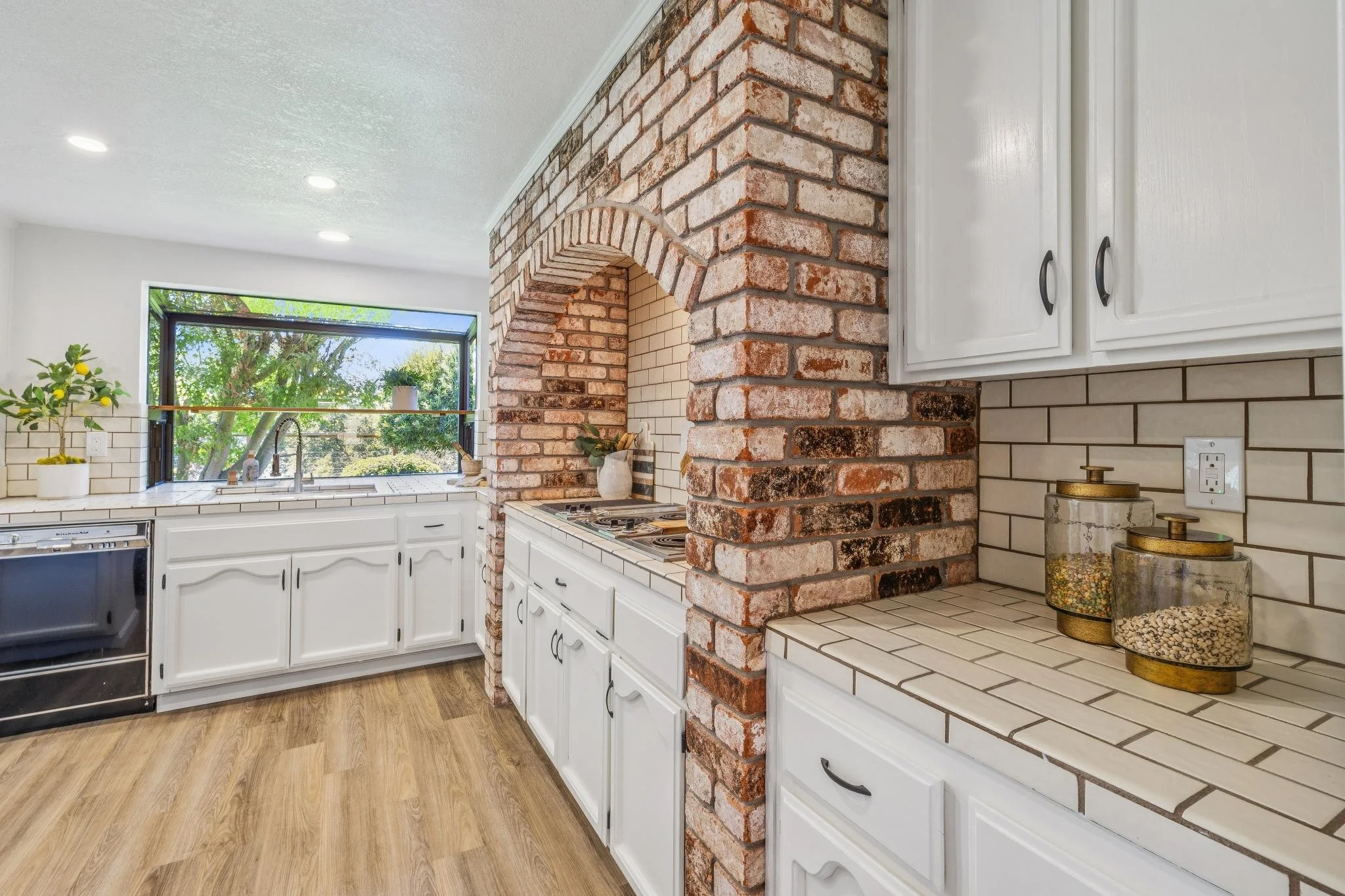Renovated kitchen with white cabinets, a brick archway, and large window overlooking greenery.