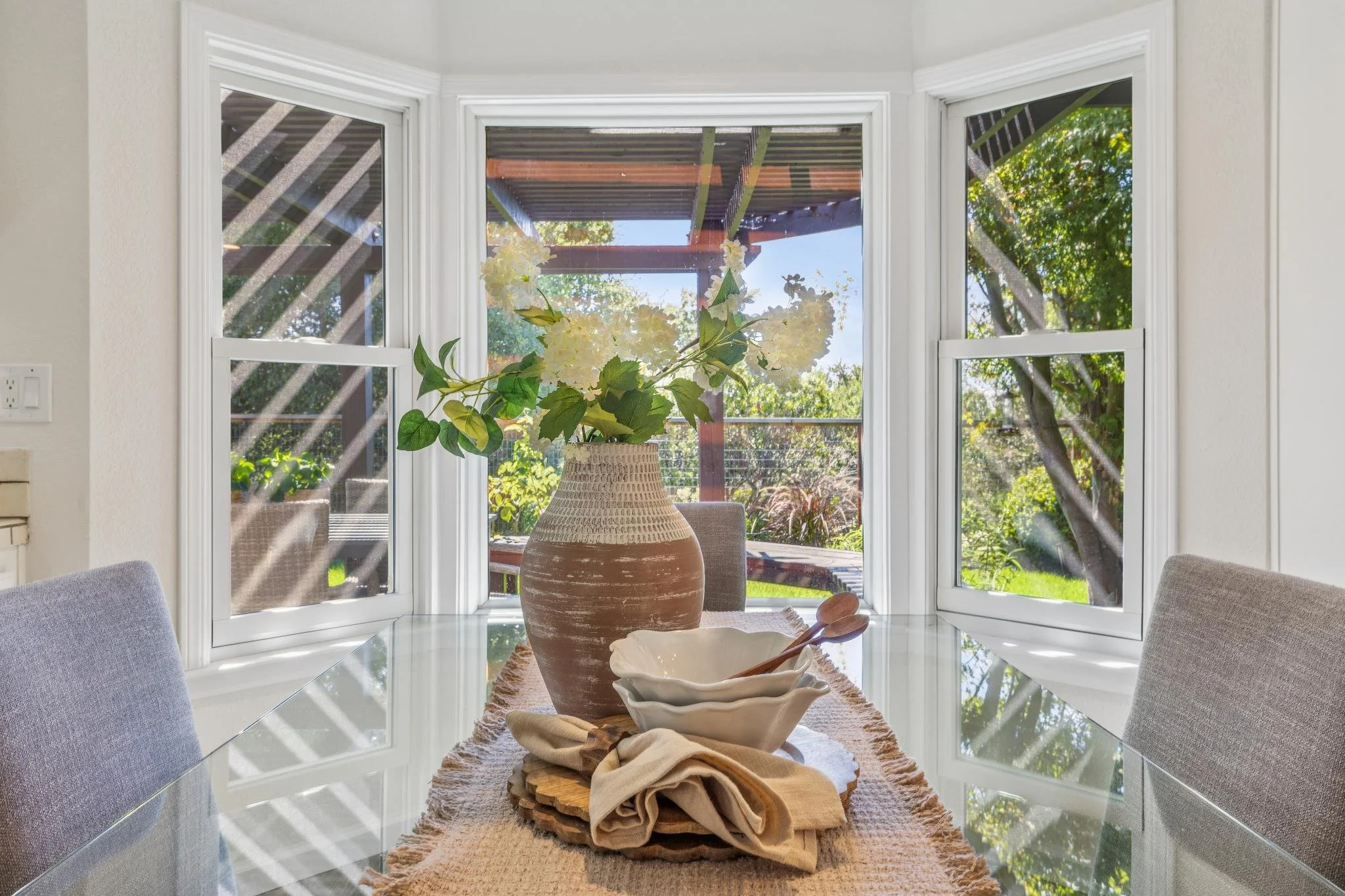 A dining area with a glass table, a vase of white flowers, and windows showing a backyard with trees and a patio.