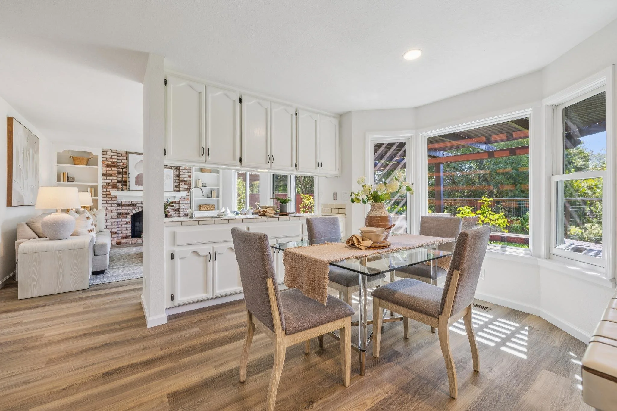 Bright dining area in a home with large windows, a glass dining table, six upholstered chairs, a wooden vase with flowers, and a view of a backyard with trees outside.