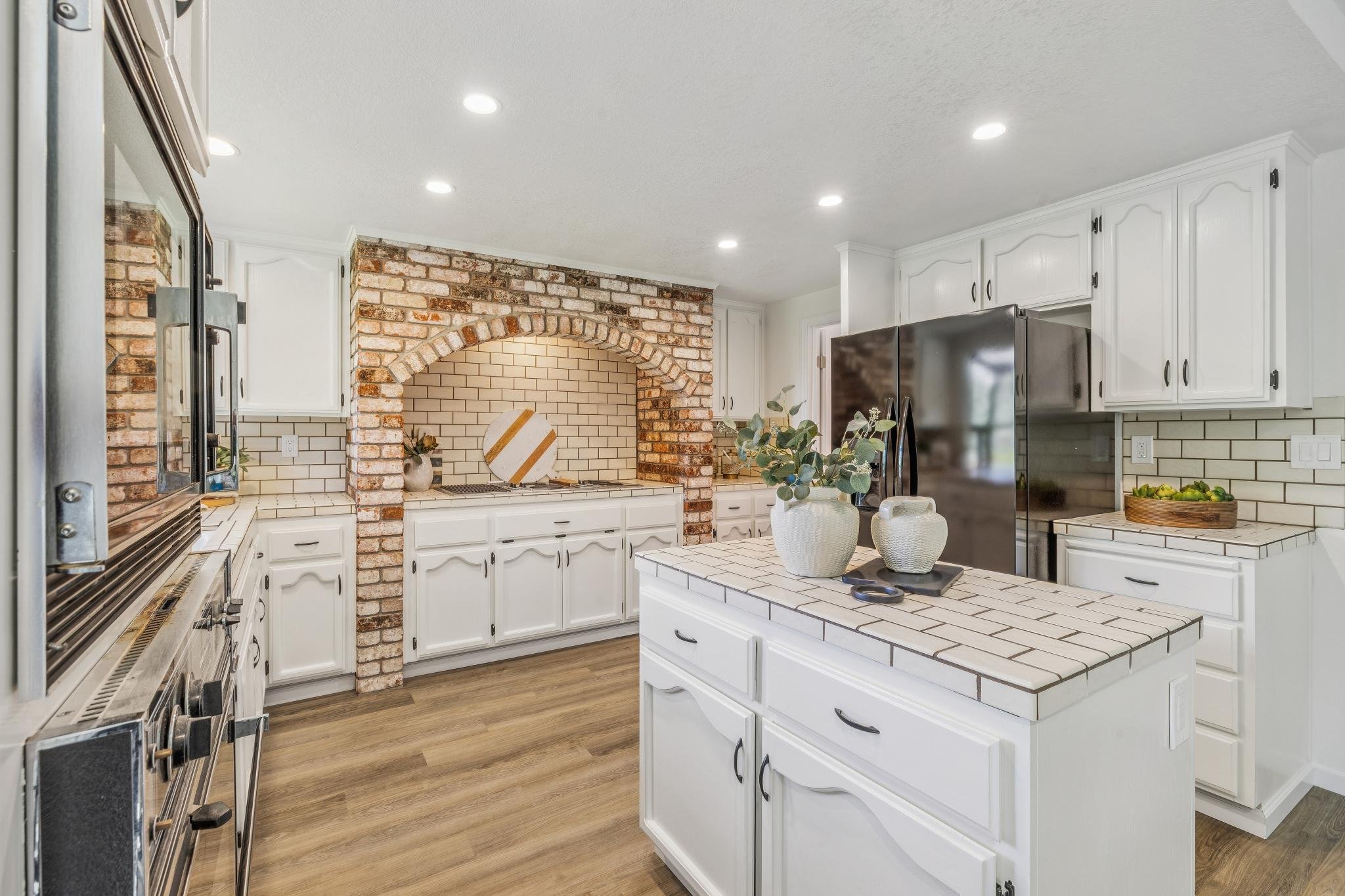 A kitchen with white cabinets and a brick arch over an oven space, a stainless steel refrigerator, a tiled kitchen island with white vases and greenery, and hardwood flooring.