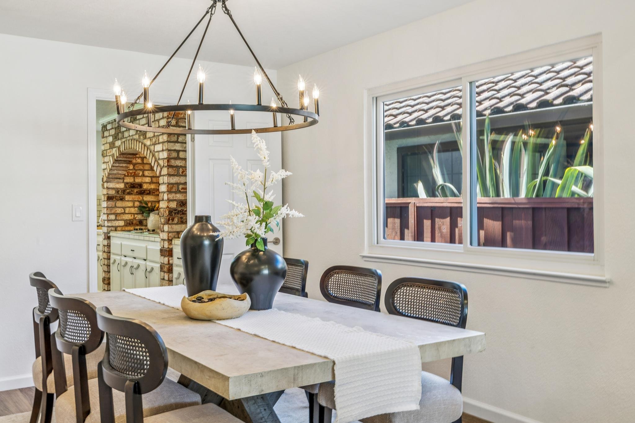 Modern dining room with a rectangular table, black chairs, tall black vases, a white runner, decorative bowl, large window showing roof tiles and greenery, chandelier with candle-like bulbs, and a brick archway leading to the kitchen.