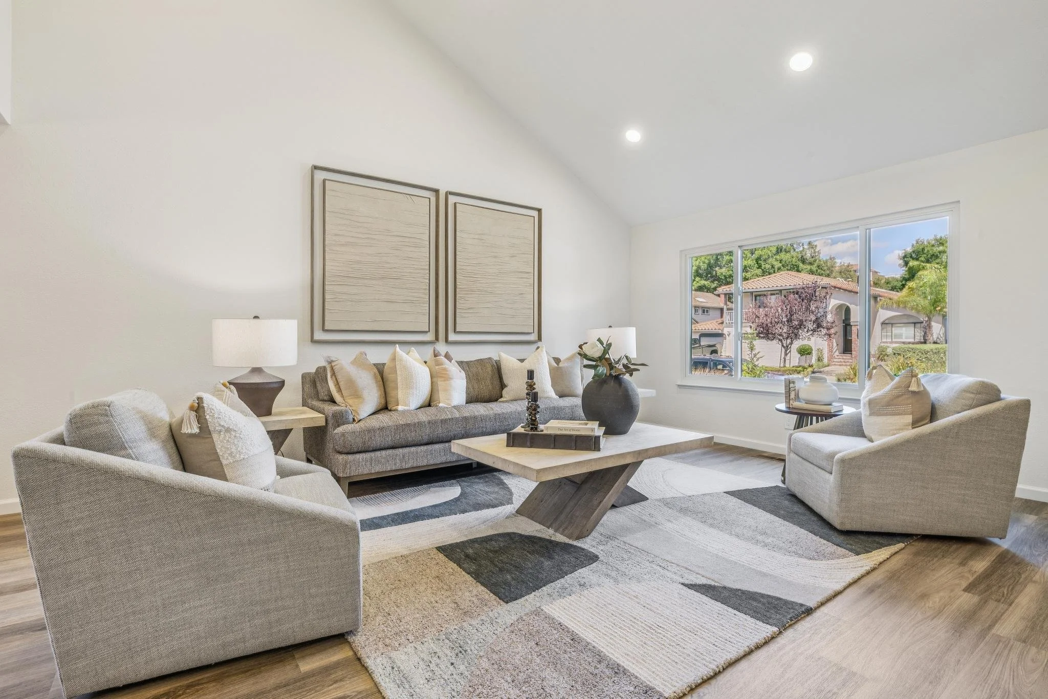 A living room with beige upholstered sofas, a wooden coffee table, off-white pillows, a large window showing houses and trees outside, and wall art above the sofa.