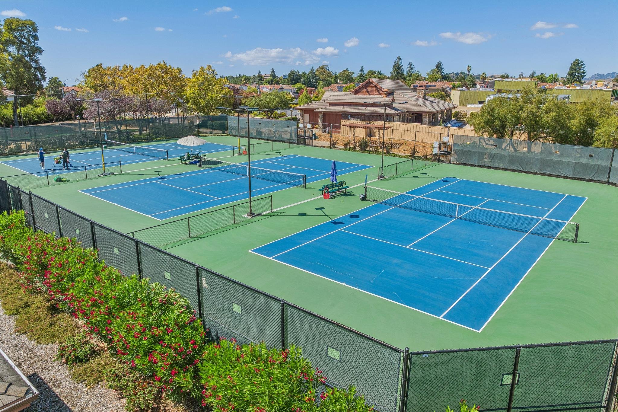 Multiple tennis courts with players and spectators, surrounded by greenery and residential houses under a blue sky with scattered clouds.