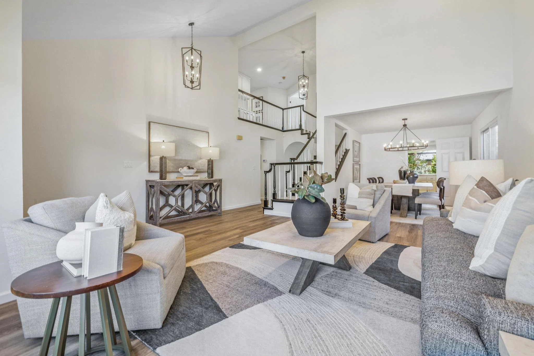 Bright, spacious living room with beige upholstered furniture, a large black vase on the coffee table, and a patterned area rug on hardwood floors. The background features a staircase with dark wood railings, pendant lights, and a dining area with a chandelier and buffet against the wall. Neutral colors dominate the decor.