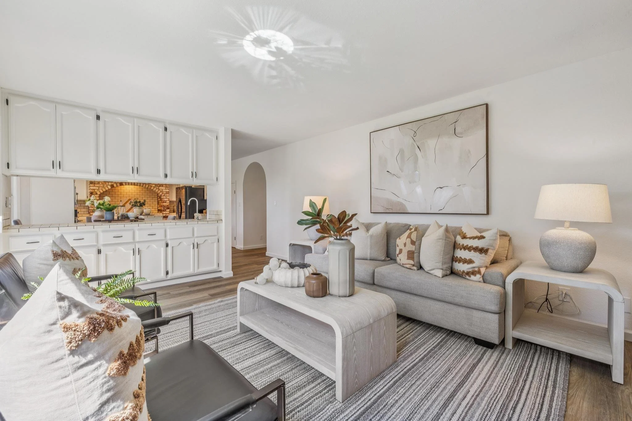 Living room with white walls, a large abstract painting, a beige sofa with patterned pillows, a coffee table with decorative items, and a nearby lamp. Part of a kitchen with white cabinets and brick arch is visible in the background.
