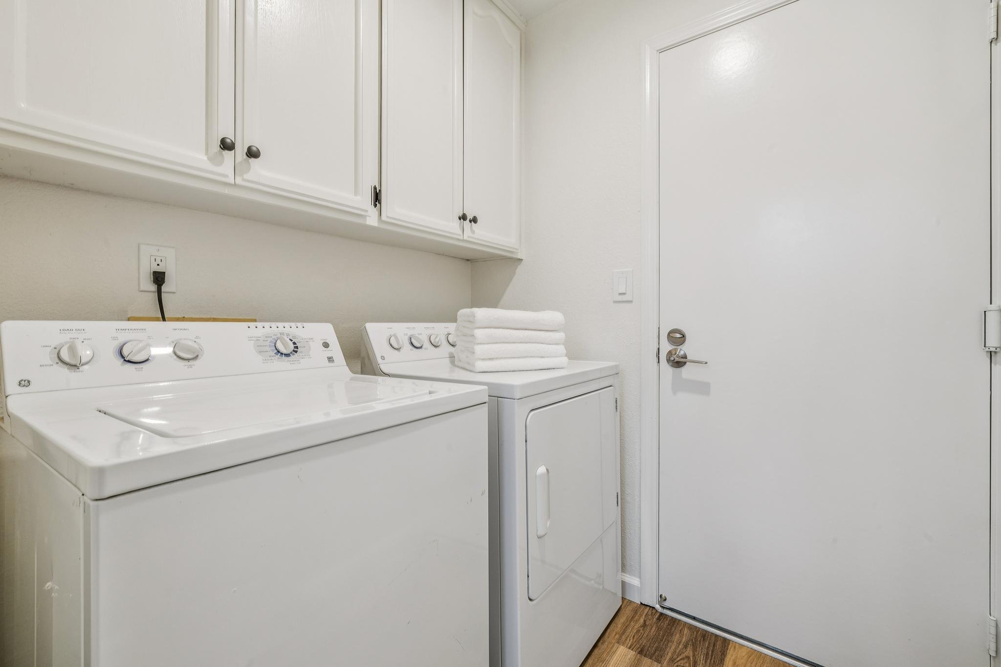 A laundry room with white cabinets, a washing machine with control knobs, a dryer, a stack of folded white towels, a wall outlet with a black plug, a white door with a round handle, a light switch, and hardwood flooring.