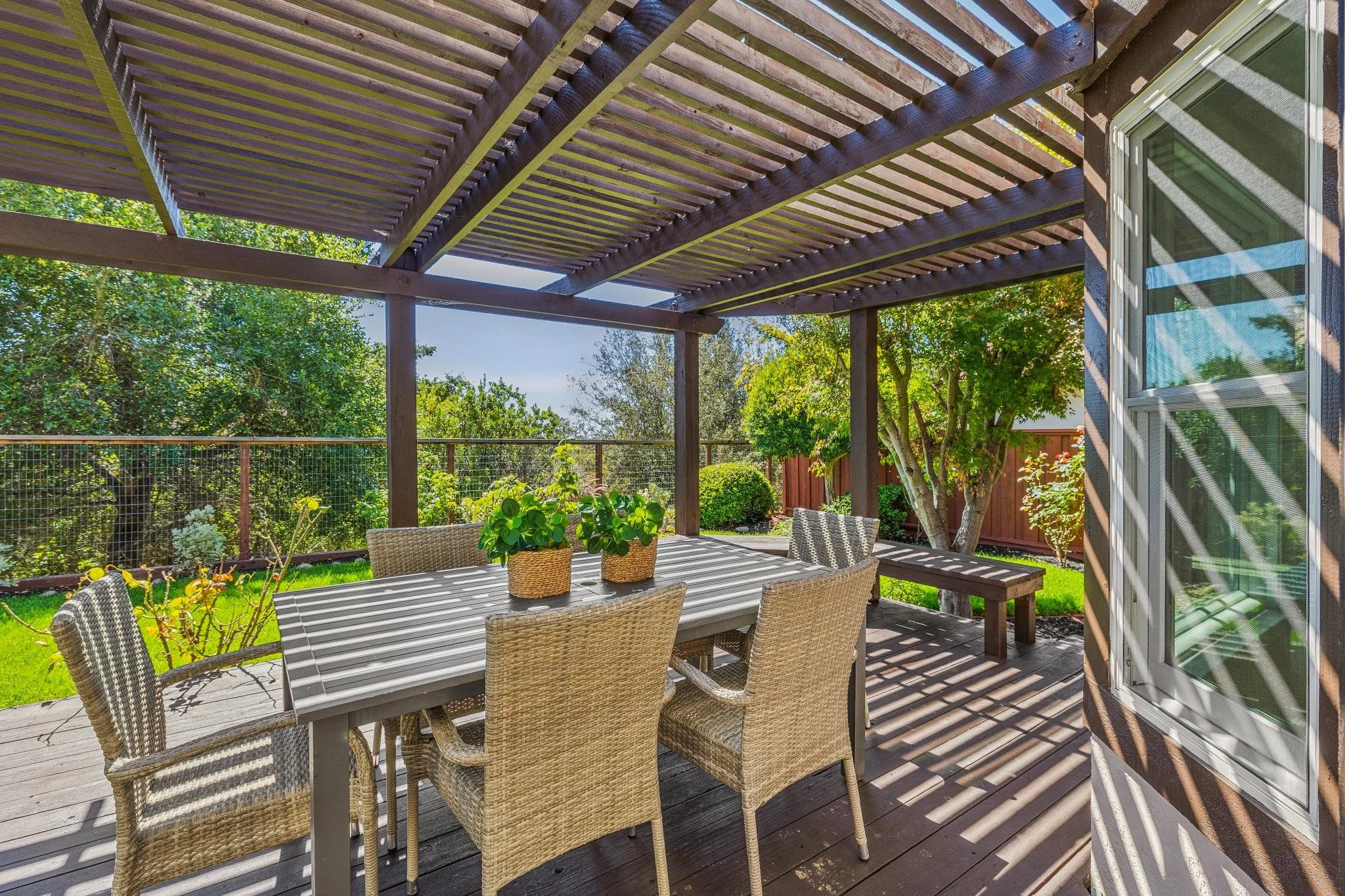 An outdoor patio area with a wooden pergola overhead, a large dining table with wicker chairs, potted plants on the table, surrounded by green trees and a wooden fence, with sunlight casting shadows.