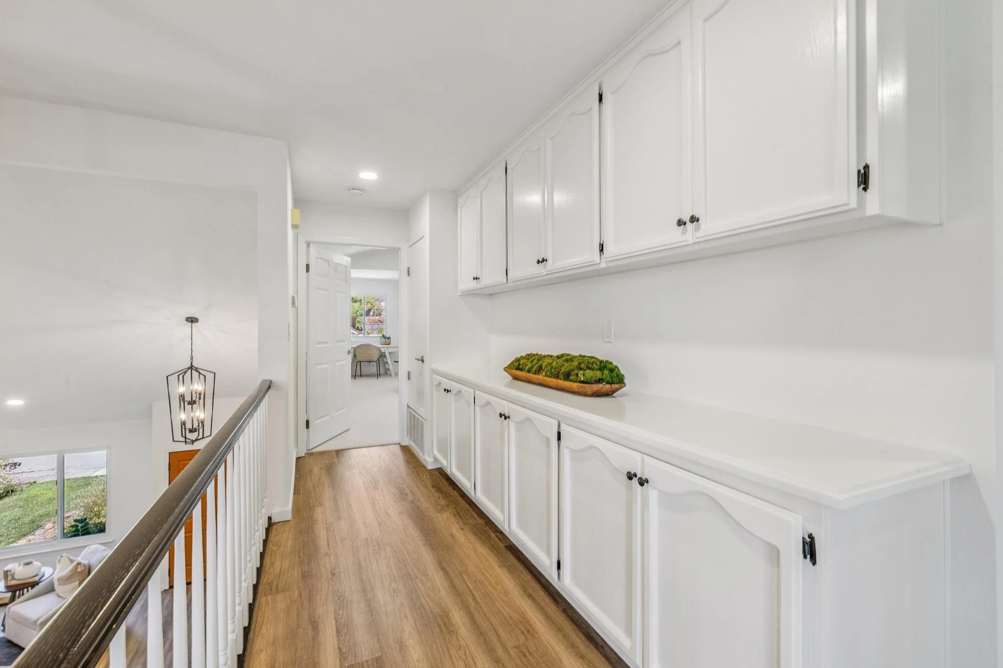 A hallway with white cabinets and a wooden floor in a home interior, with stairs and a chandelier visible at the bottom and a window at the end of the hallway.