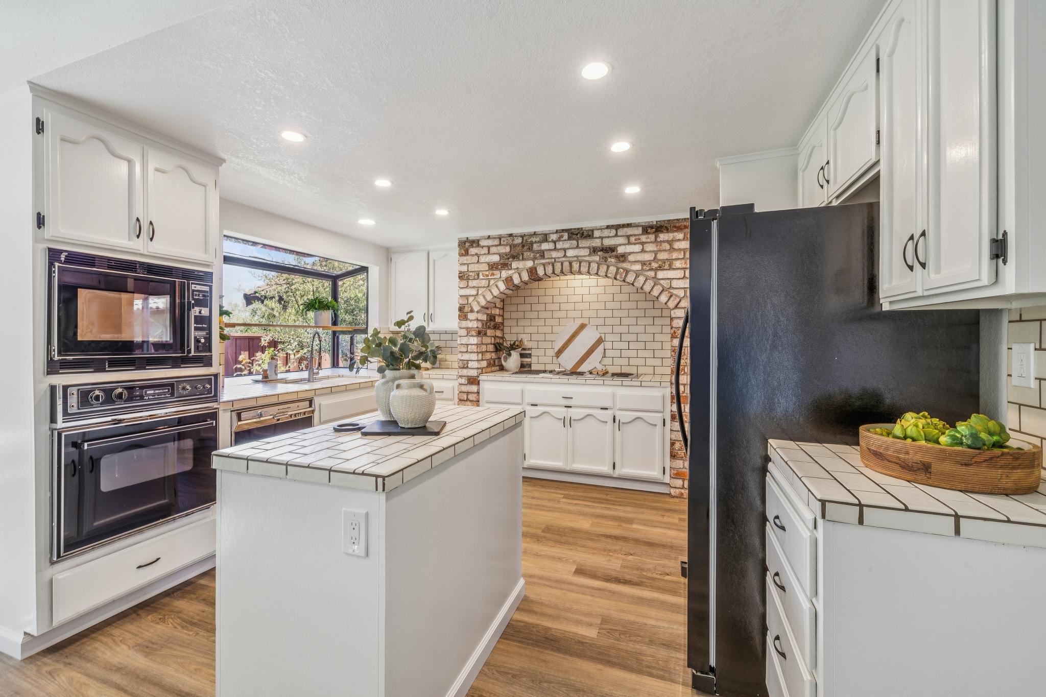 Modern kitchen with white cabinets, brick arch wall, black refrigerator, and wood flooring, featuring a window with view of trees.
