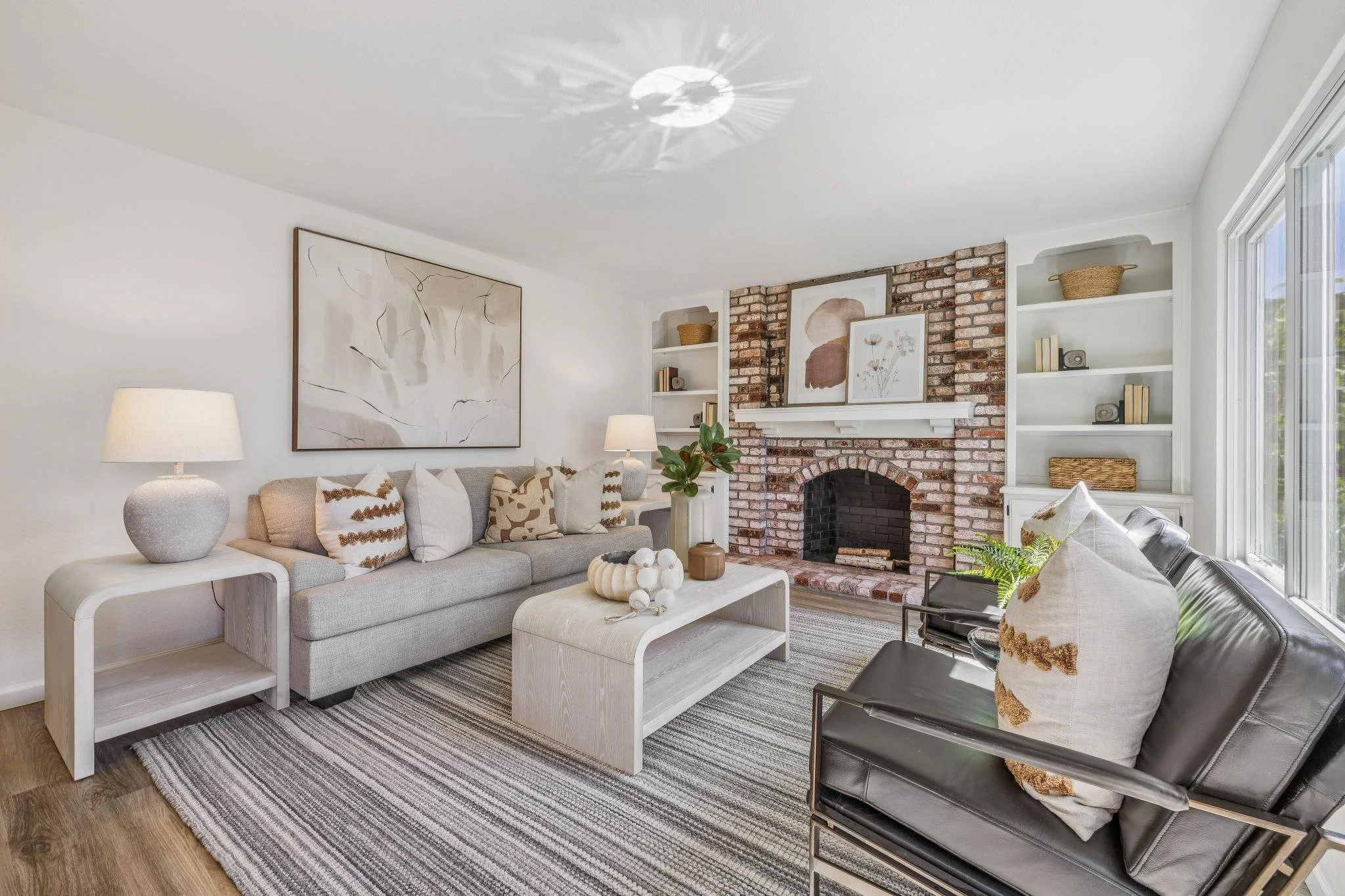 Living room with a beige sofa, black leather chair, fireplace with brick surround, white built-in shelves, abstract art, potted plant, lamp, and decorative items.