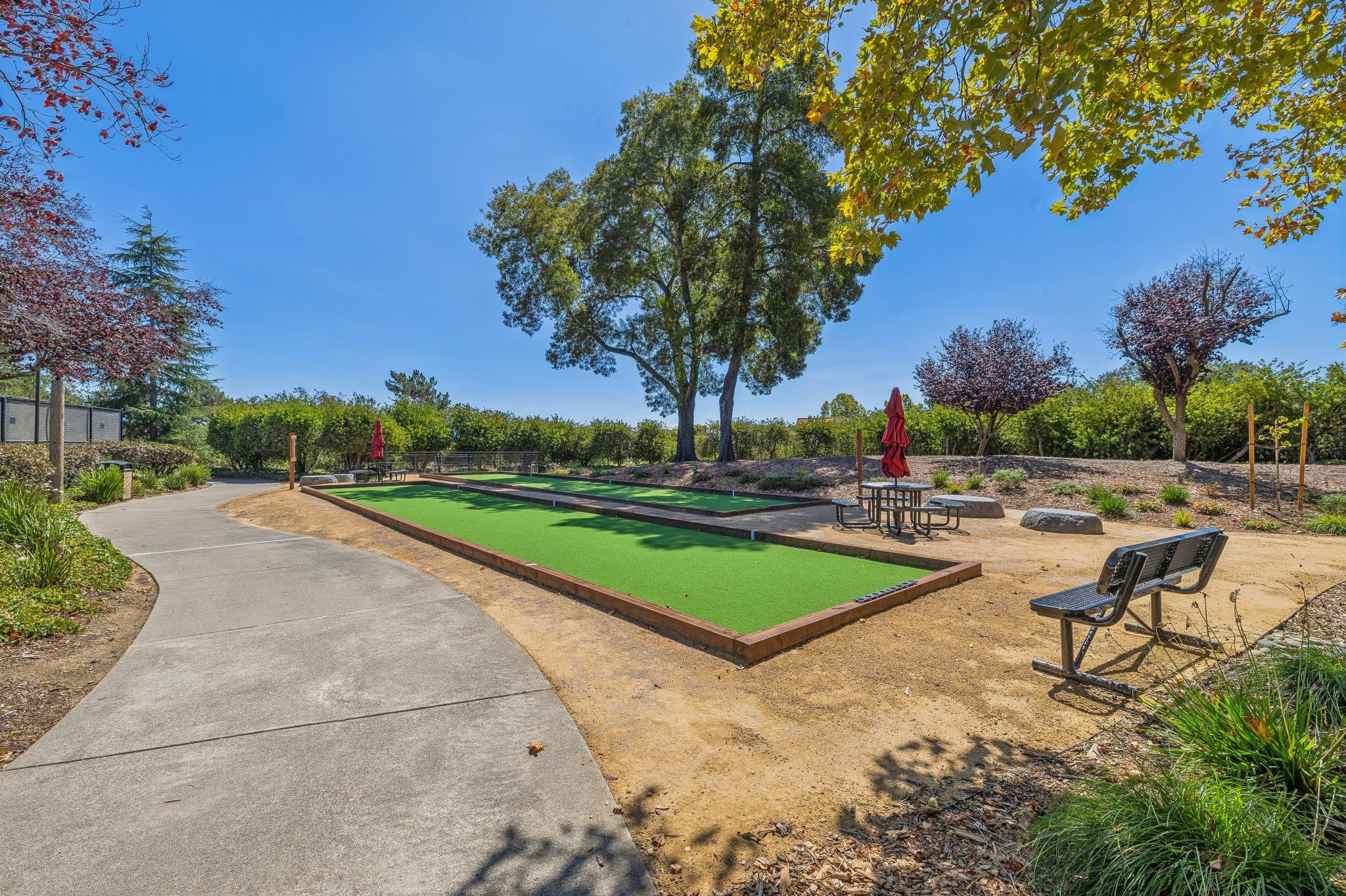 A park with a paved walking path, a patch of green artificial turf, surrounding trees with green and reddish leaves, benches, and red umbrellas under a clear blue sky.