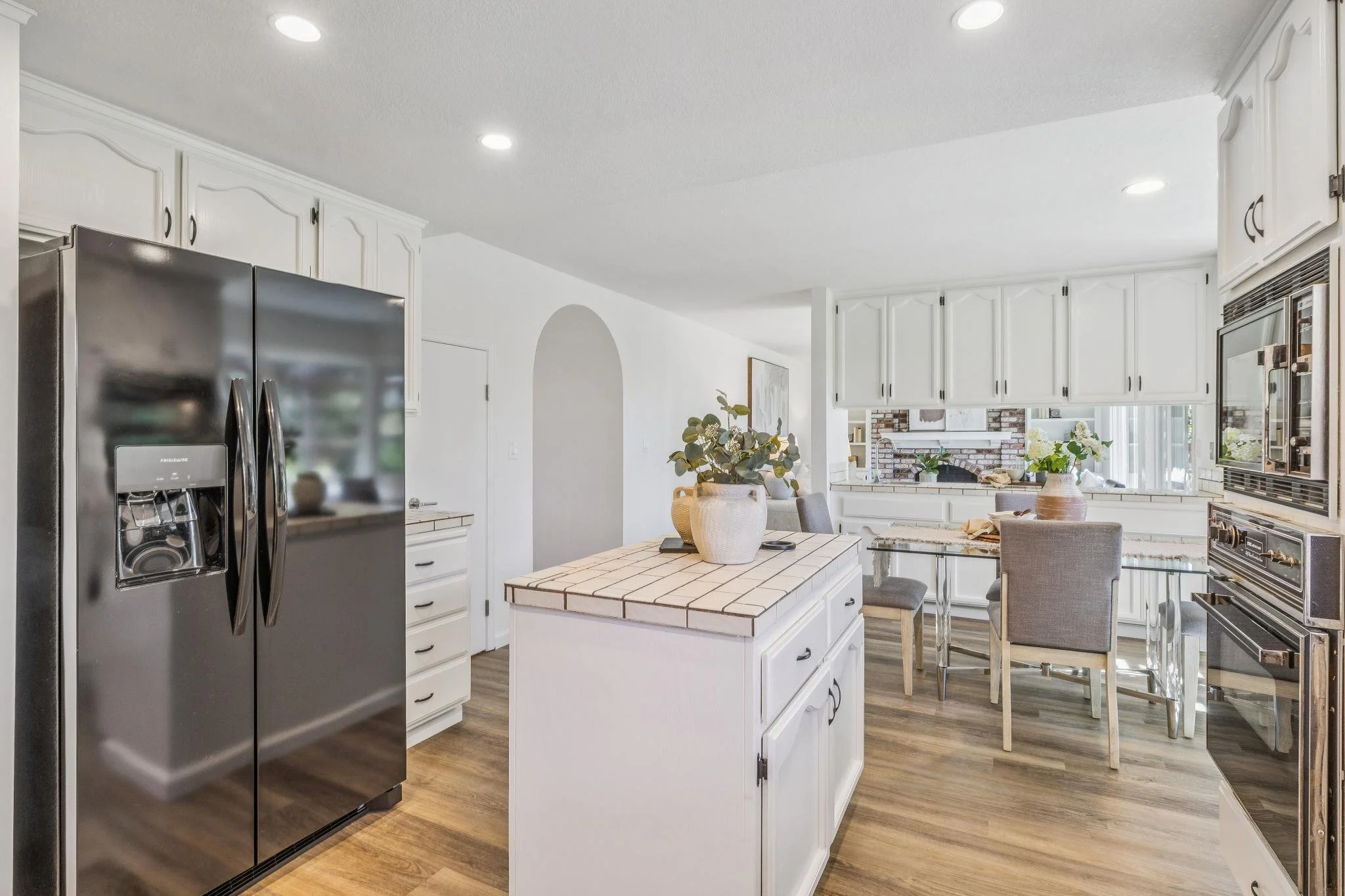 Bright kitchen with white cabinets, stainless steel refrigerator, small island with tiled top, and a dining area with a table and gray chairs. Vases with green plants on the island and in the background near a fireplace.